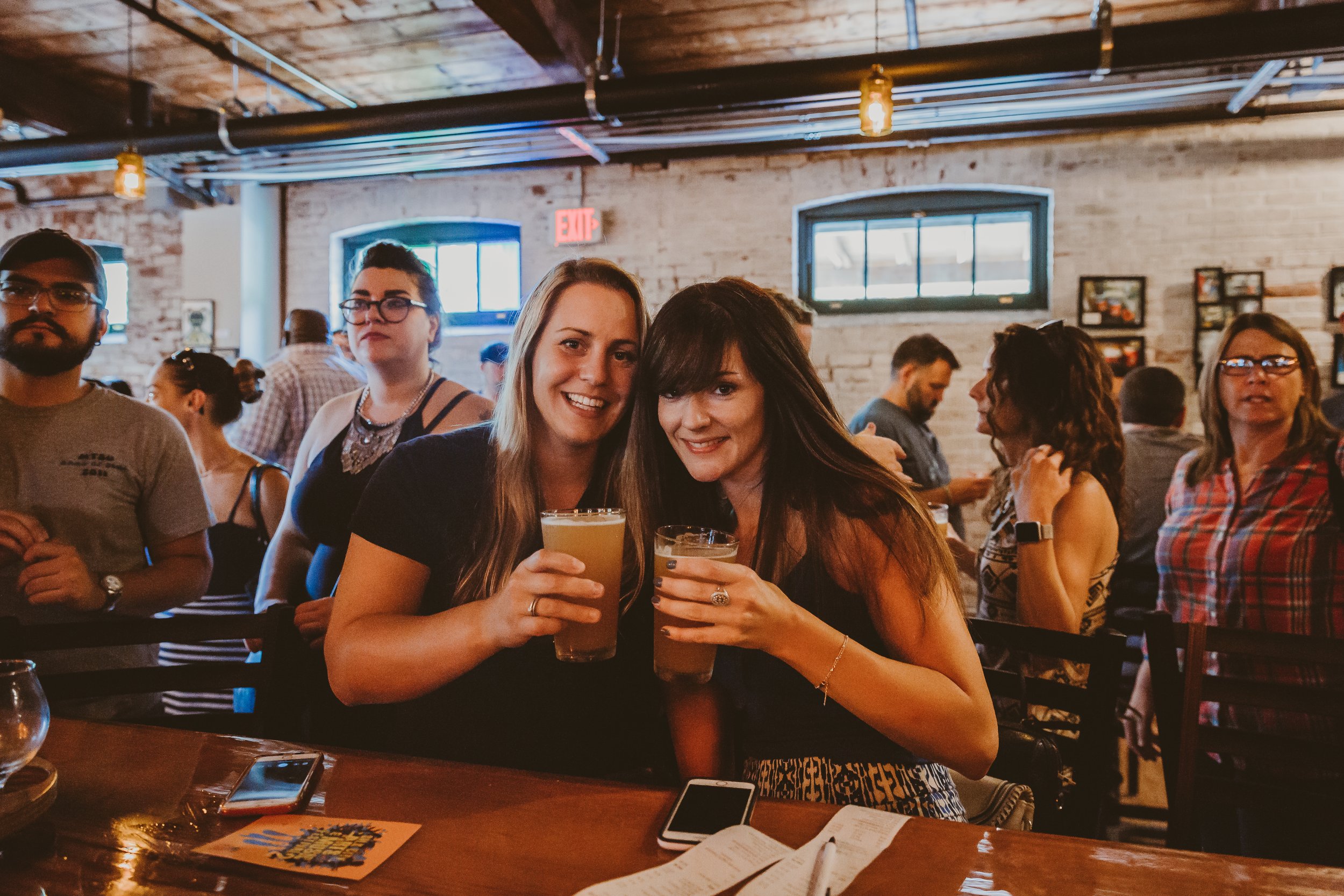 Two women smiling and holding drinks at a bar or restaurant with a crowd of people in the background.