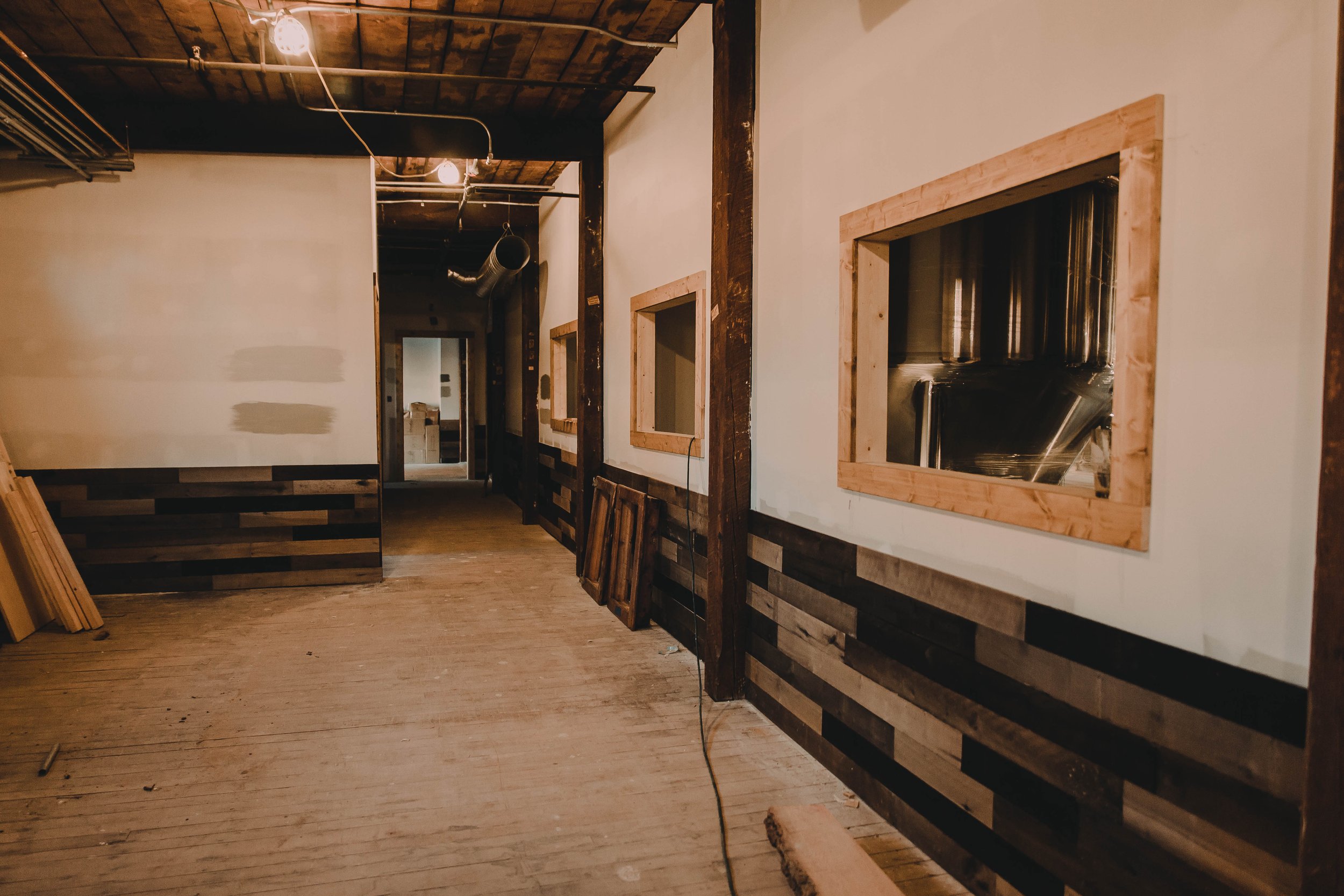 Interior of a building under construction with unfinished wooden accents, exposed pipework, and a hardwood floor.