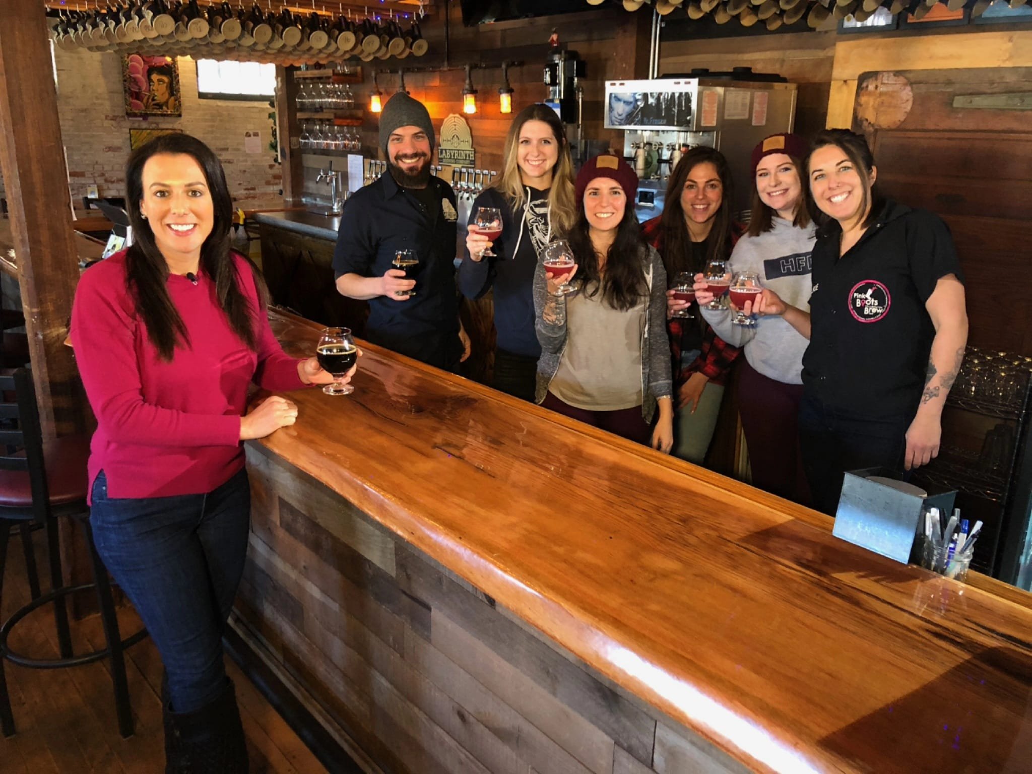 A group of eight people, mostly women, standing behind a wooden bar counter in a bar or brewery, holding glasses of dark beverages and smiling at the camera.