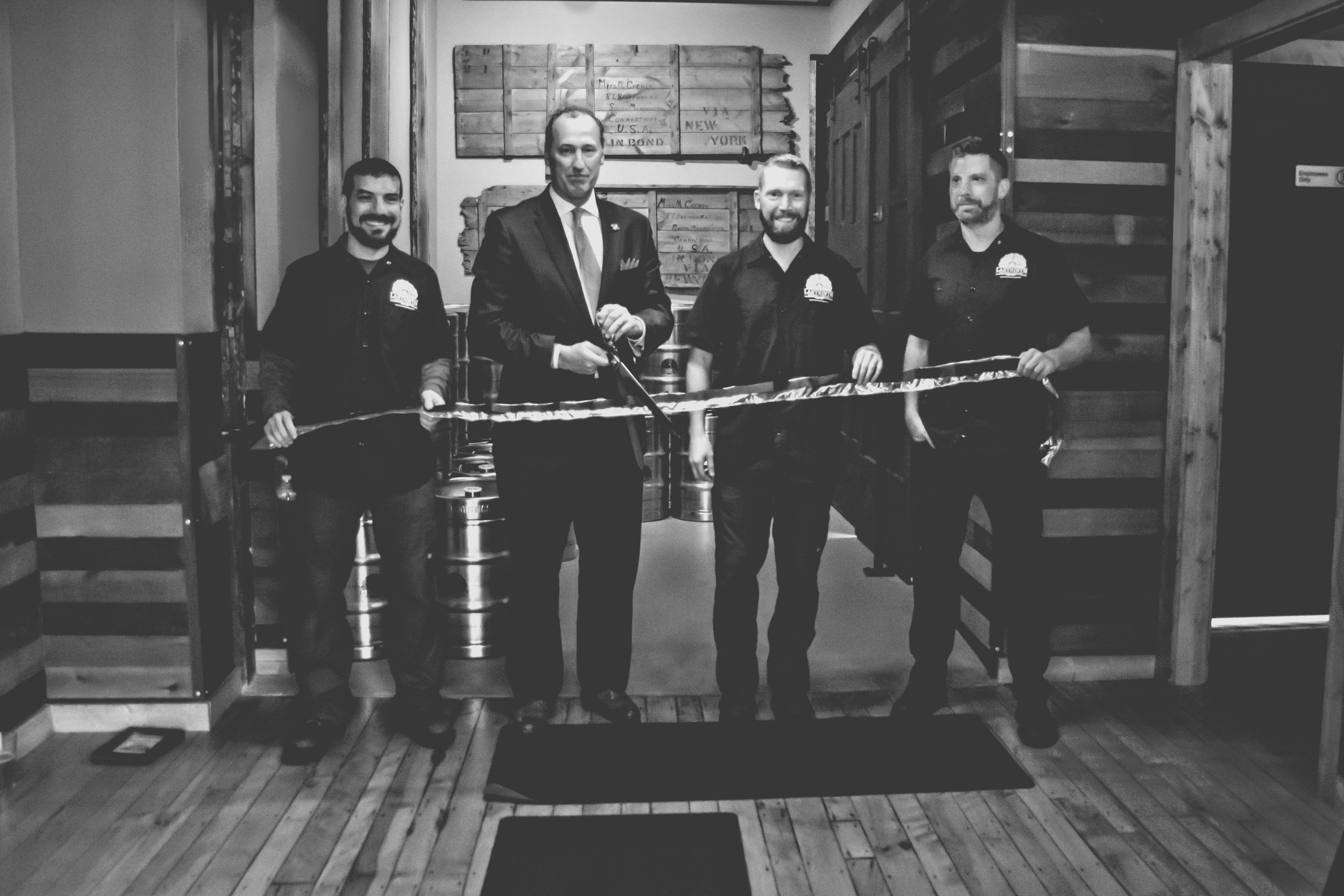 Four men standing indoors holding a long ribbon, with kegs in the background. One man in a suit and three in matching black shirts. It appears to be a ribbon-cutting event.