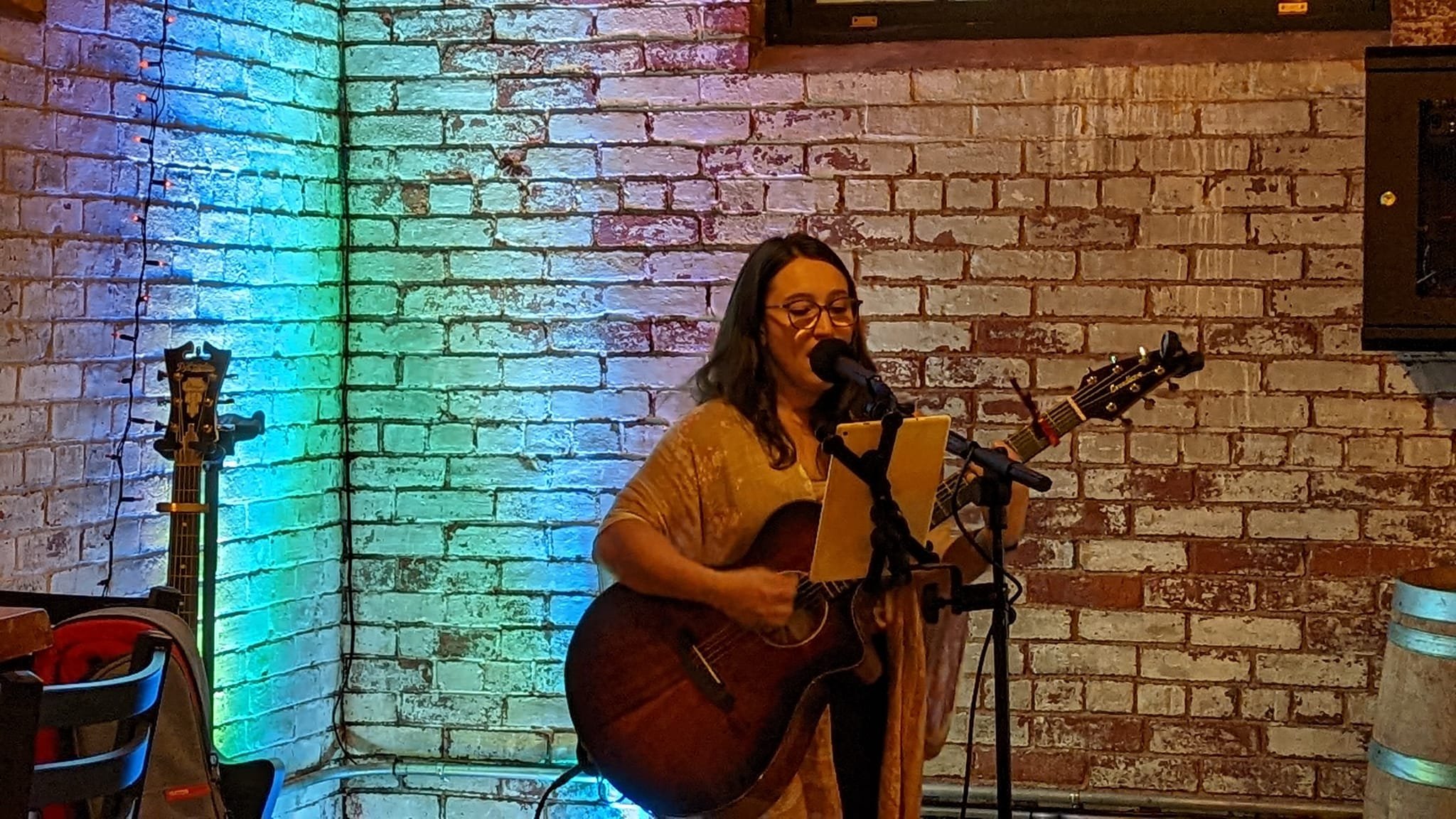 A woman wearing glasses and a tan shirt performs with an acoustic guitar and microphone in front of a brick wall illuminated with colorful lights.