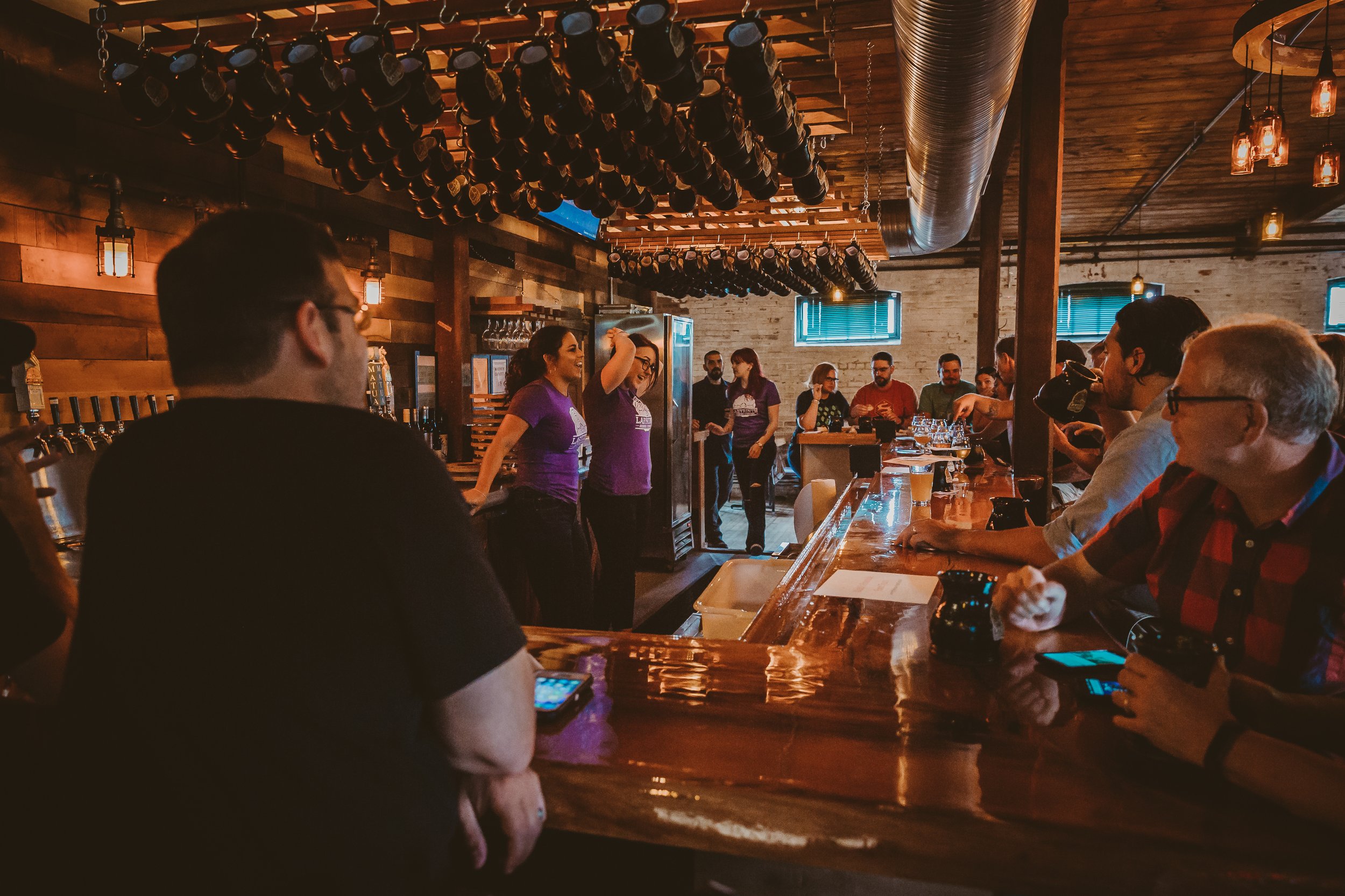 A lively bar scene with patrons sitting at the counter and two women in purple shirts talking to the crowd, with bottles hanging from the ceiling.