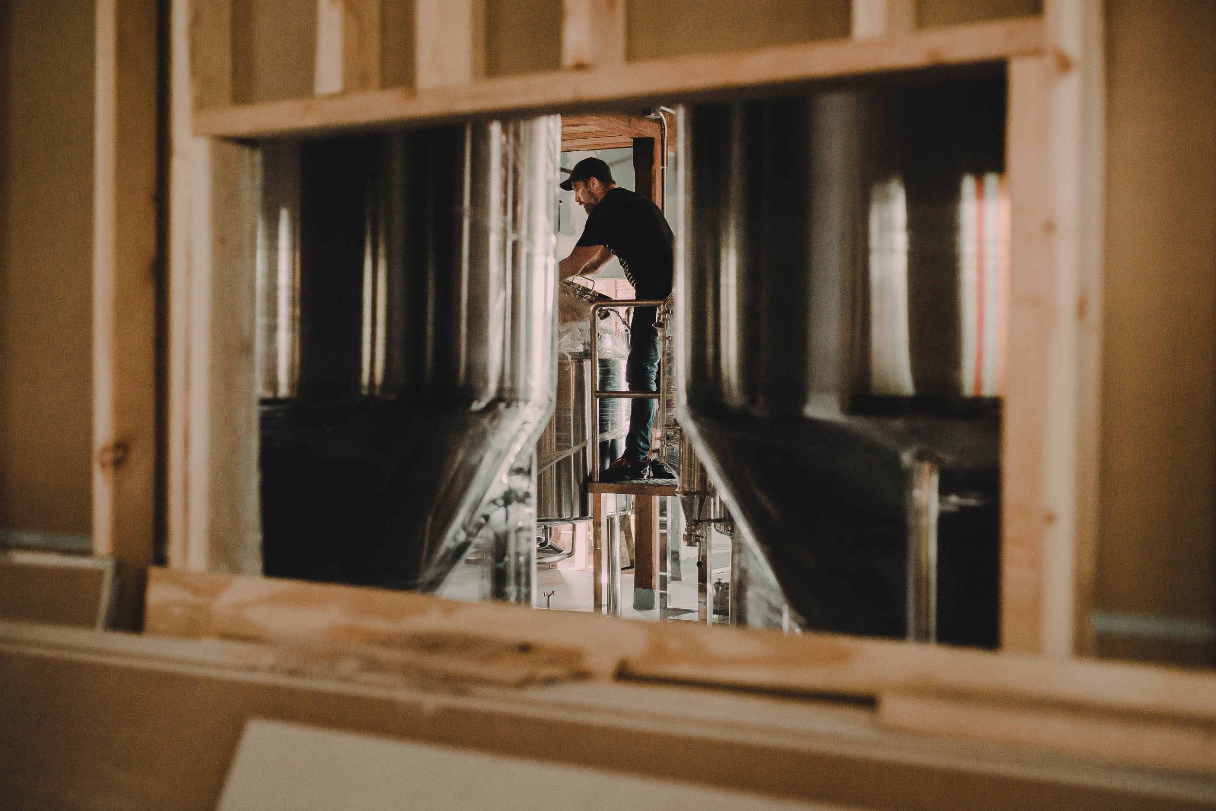 A man in a black shirt and jeans standing on a small platform working on plumbing pipes framed through a rectangular opening in a wall under construction.
