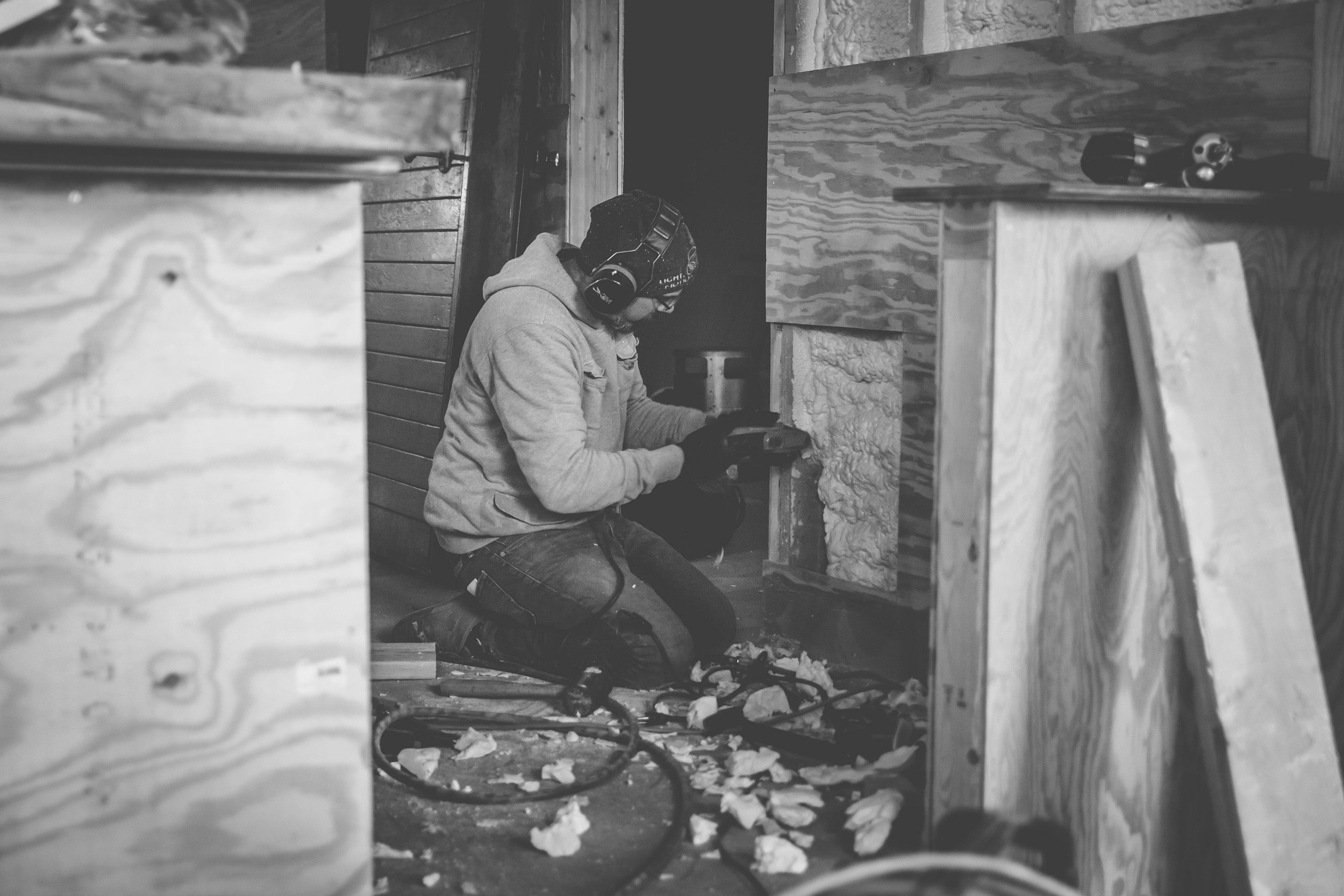 A person wearing a helmet and gloves working on a woodworking project indoors, kneeling on the floor surrounded by wood scraps and tools.
