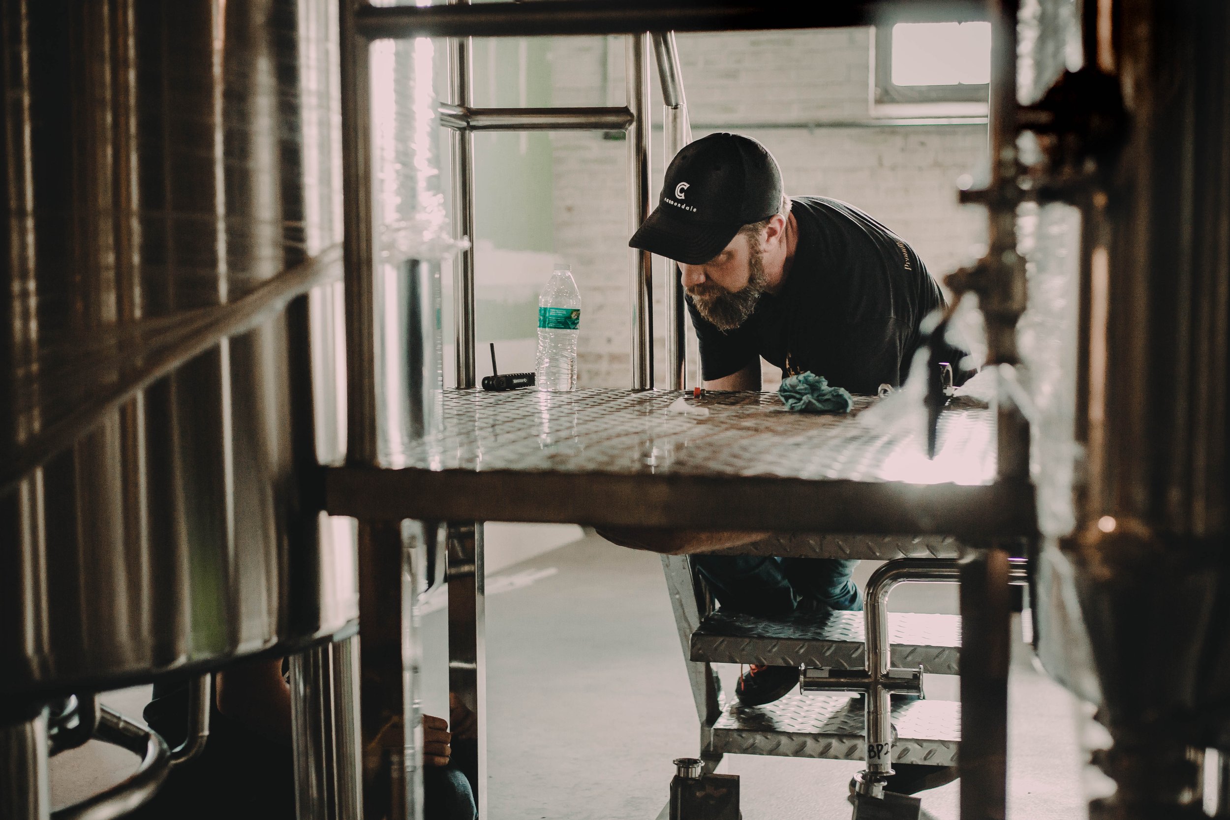 A man with a beard and cap working on a tank or stainless steel structure inside a building, with a water bottle, rag, and tools on the surface.