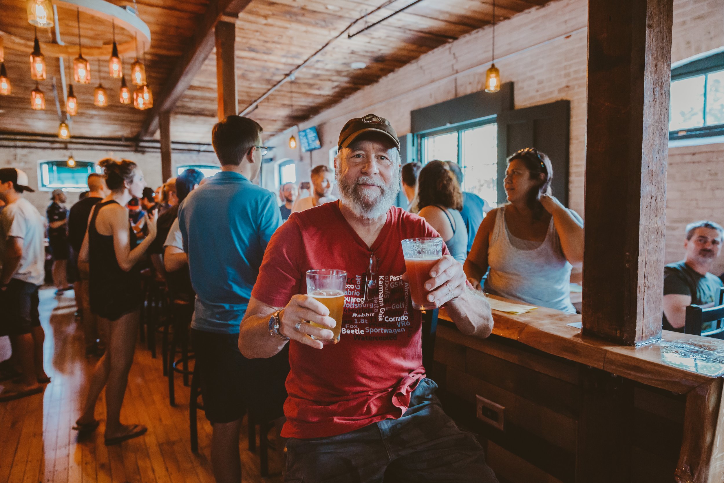 A smiling elderly man with a beard wearing a red T-shirt and a black cap sitting at a bar holding two beers in a lively, rustic bar environment with many people socializing.