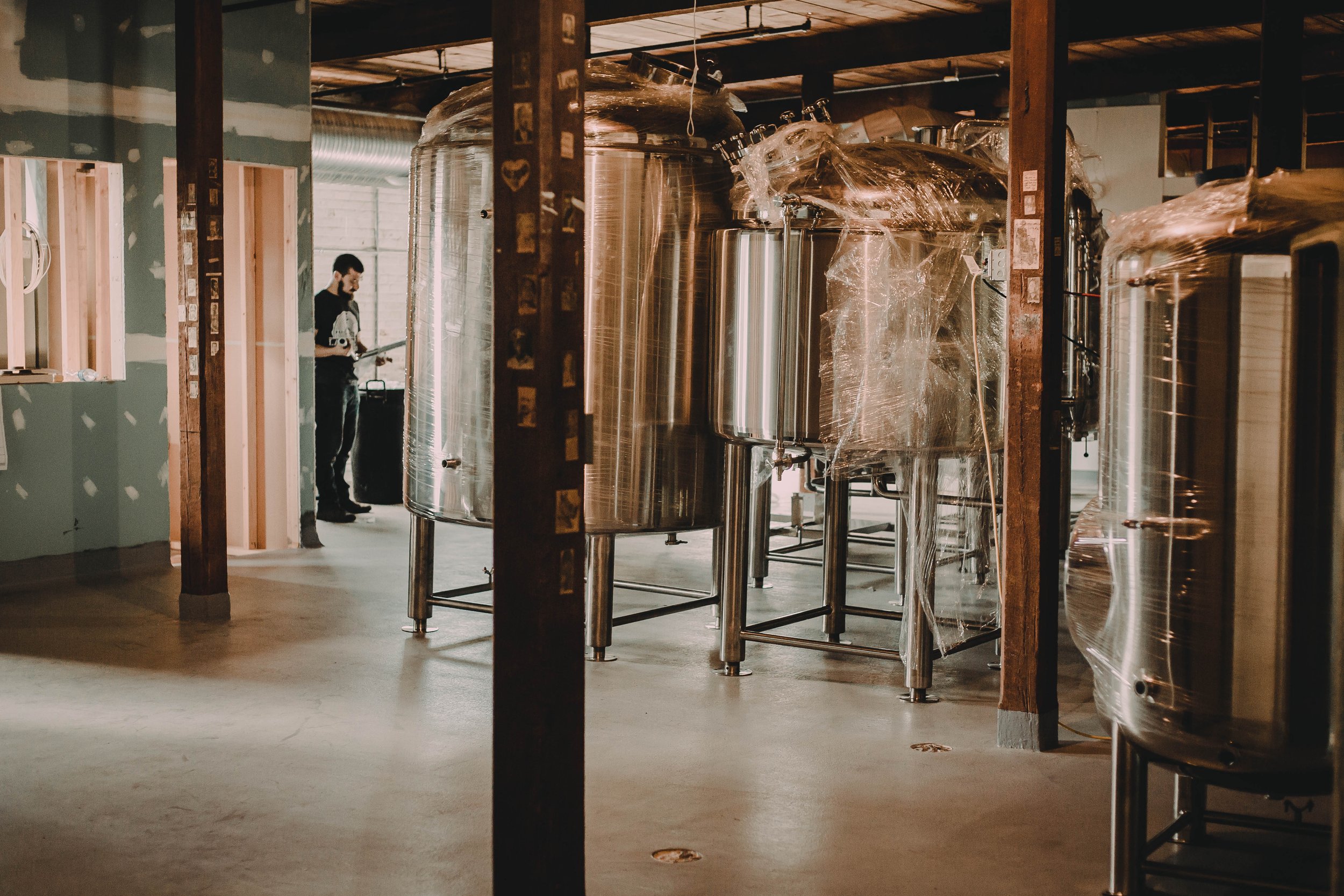 Industrial brewery or winery interior with stainless steel tanks, a man standing in the background, and unfinished walls.
