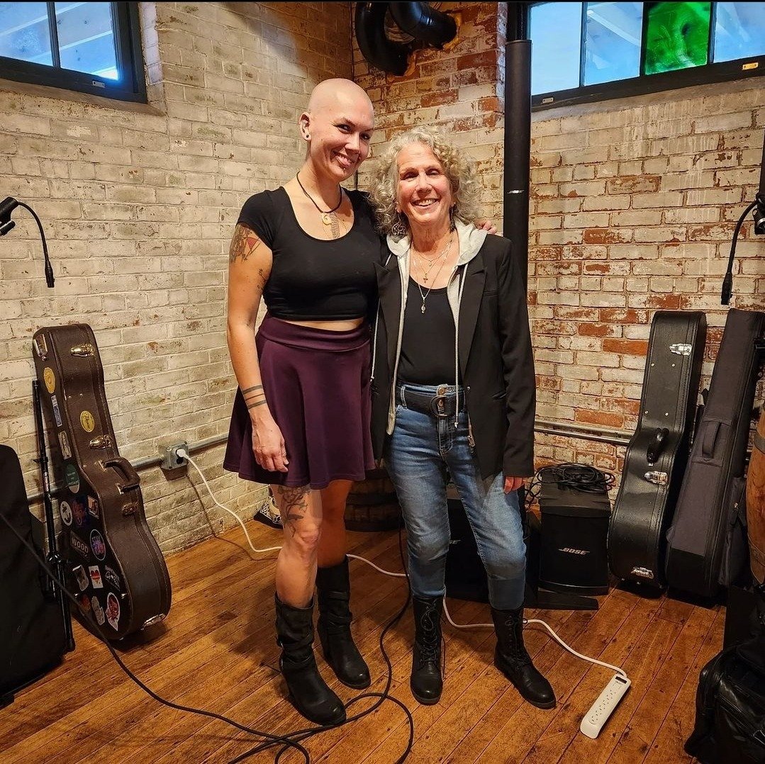 Two women standing next to each other, smiling, in a music studio with brick walls. One woman has a shaved head and tattoos, wearing a black crop top, purple skirt, and black boots. The other woman has curly gray hair, wearing a black jacket, black s