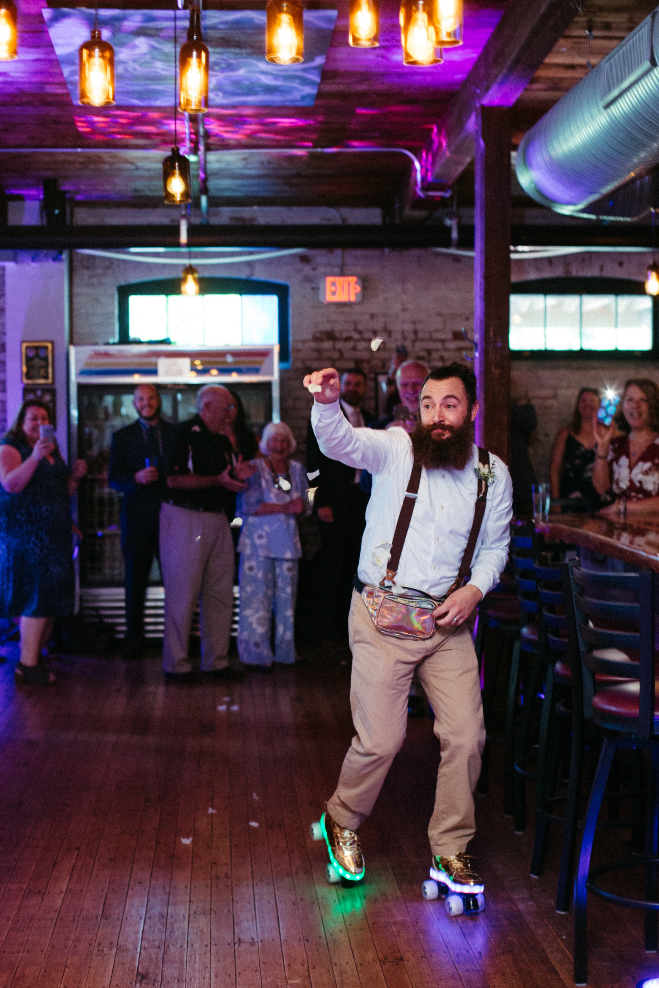 A man with a beard riding a roller skate with lights through a party or celebration with people clapping and cheering in the background, under warm hanging lights.