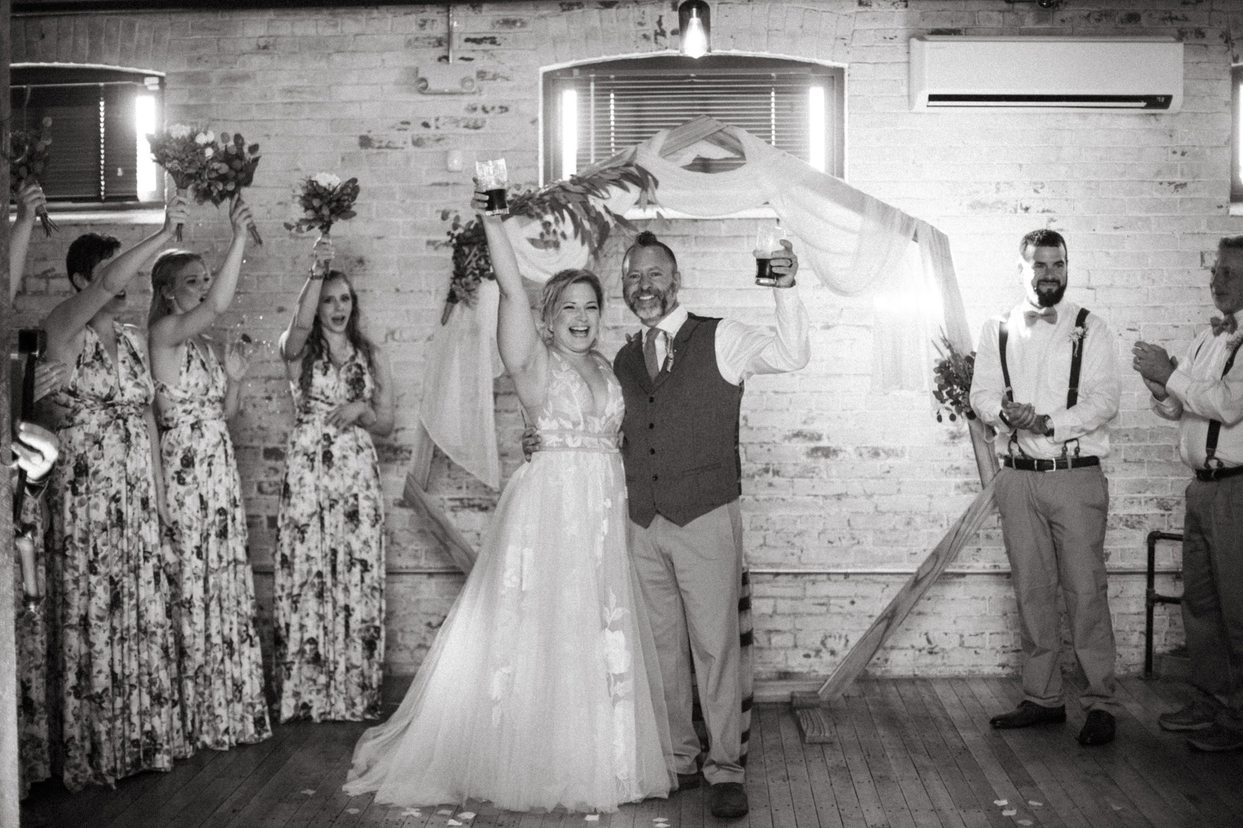 Black and white wedding photo of a bride and groom celebrating with friends and family in a rustic indoor setting.