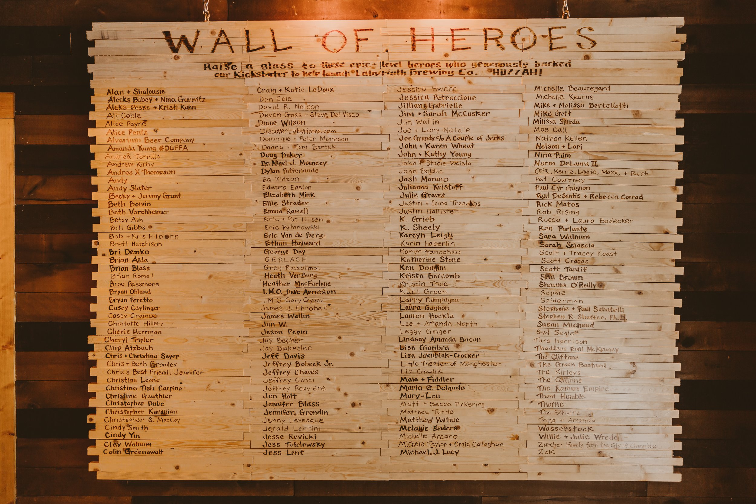 Wooden wall with a large sign titled 'Wall of Heroes' listing names of individuals who donated to a Kickstarter campaign, organized in columns, with some names highlighted in orange.