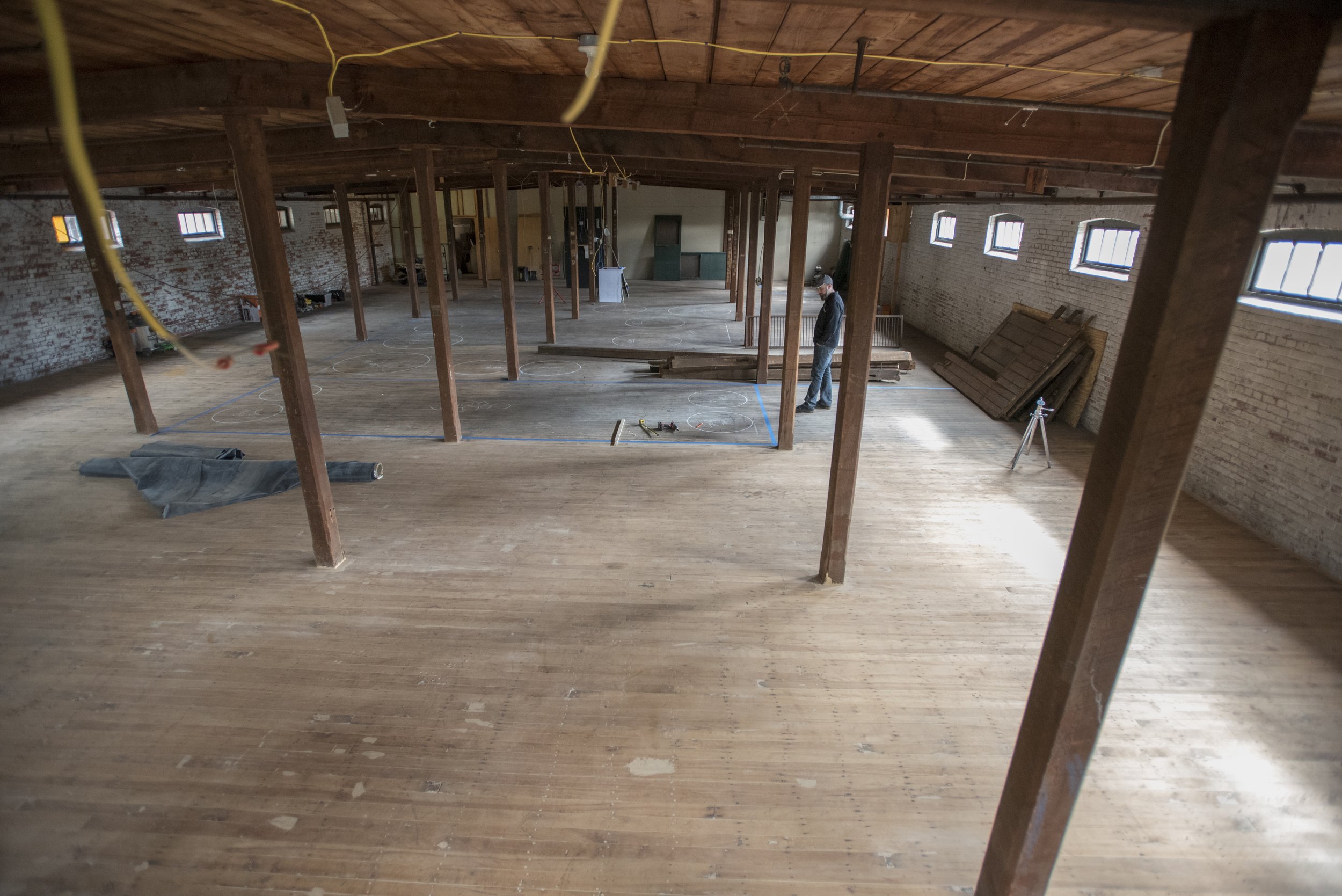 Interior of a large, open-concept space under construction with exposed brick walls, wooden floors, and wooden support beams. A man is standing near the back, and various tools and construction materials are scattered around.