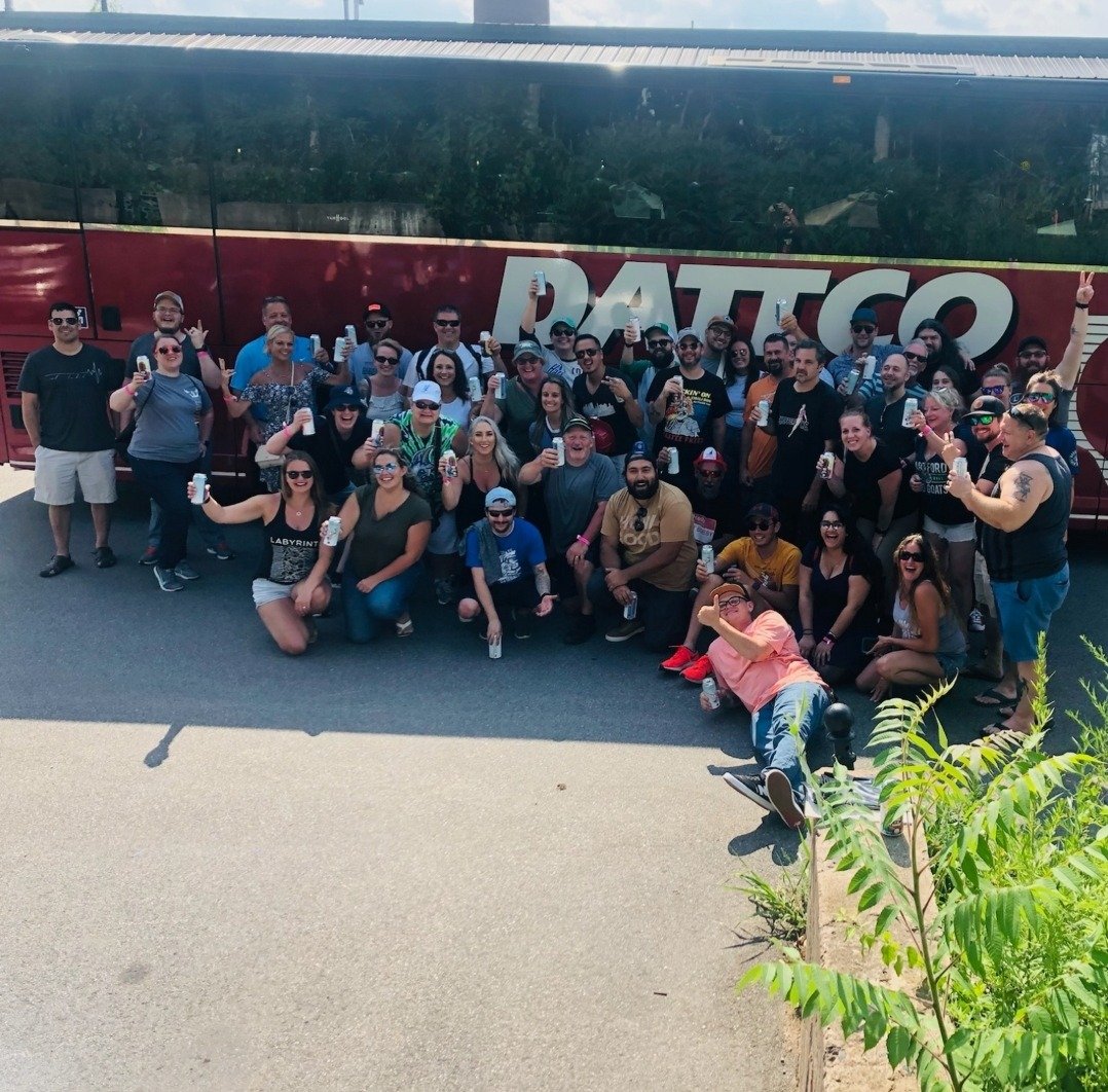 Large group of people posing for a photo in front of a bus with the word 'PATTO' on it. Many are holding drinks, some are making peace signs, and the group appears happy and celebratory.