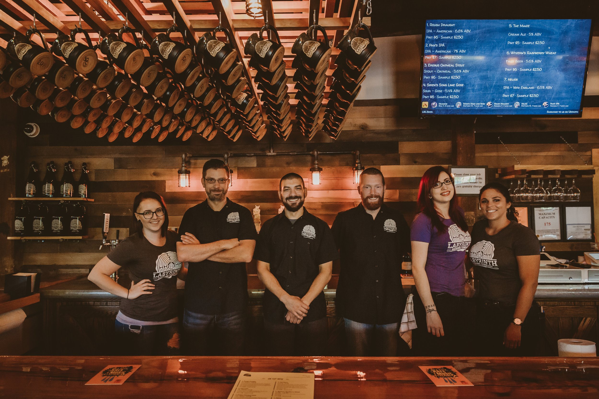 Group of five people, three men and two women, standing behind a wooden bar in a brewery or pub, with a wall of beer bottles hanging above them and a digital menu screen to the right.