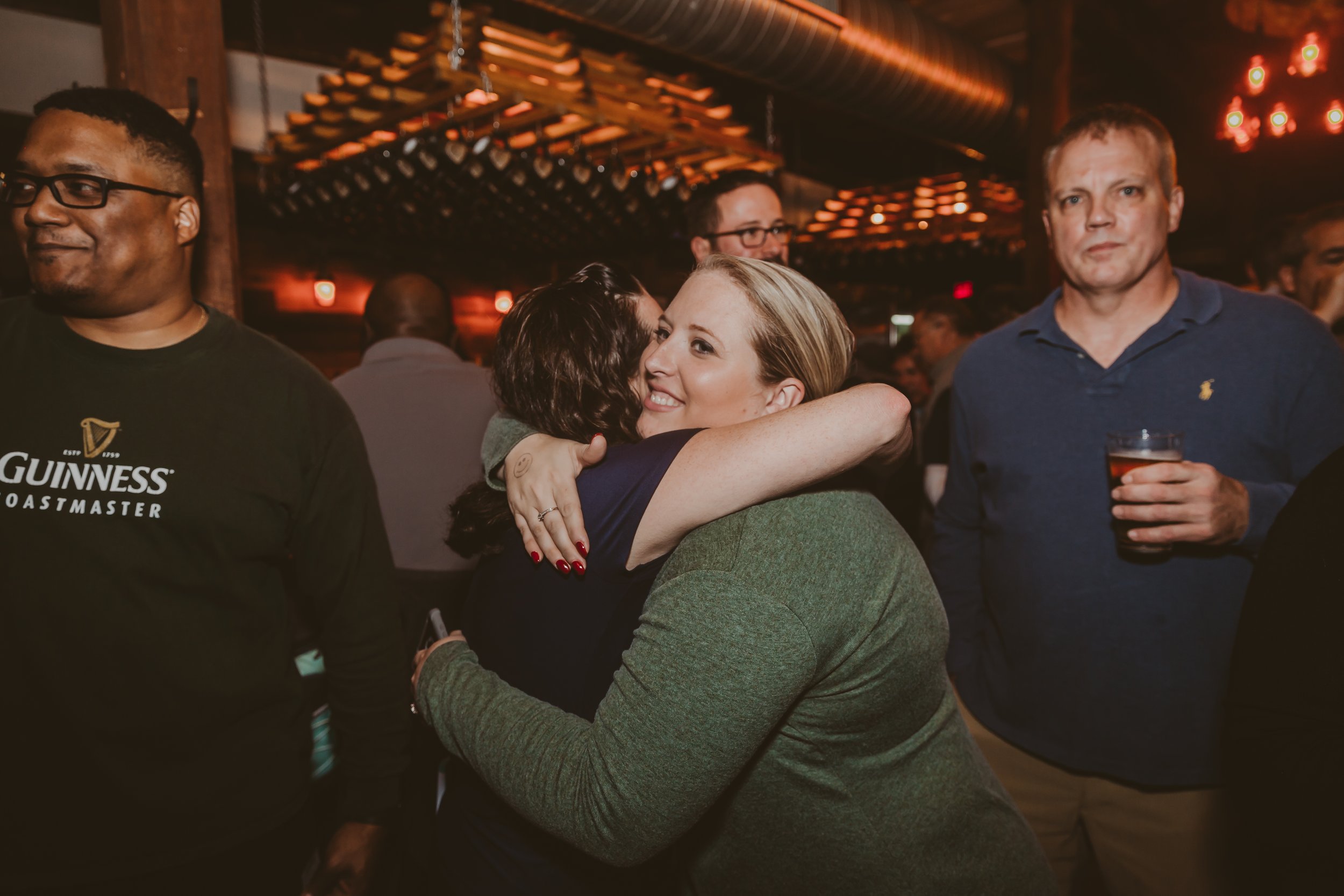 Two women hugging at a social gathering, one woman has blonde hair and is smiling, the other woman has dark hair. Several other people are visible in the background, some holding drinks in a warmly lit setting.