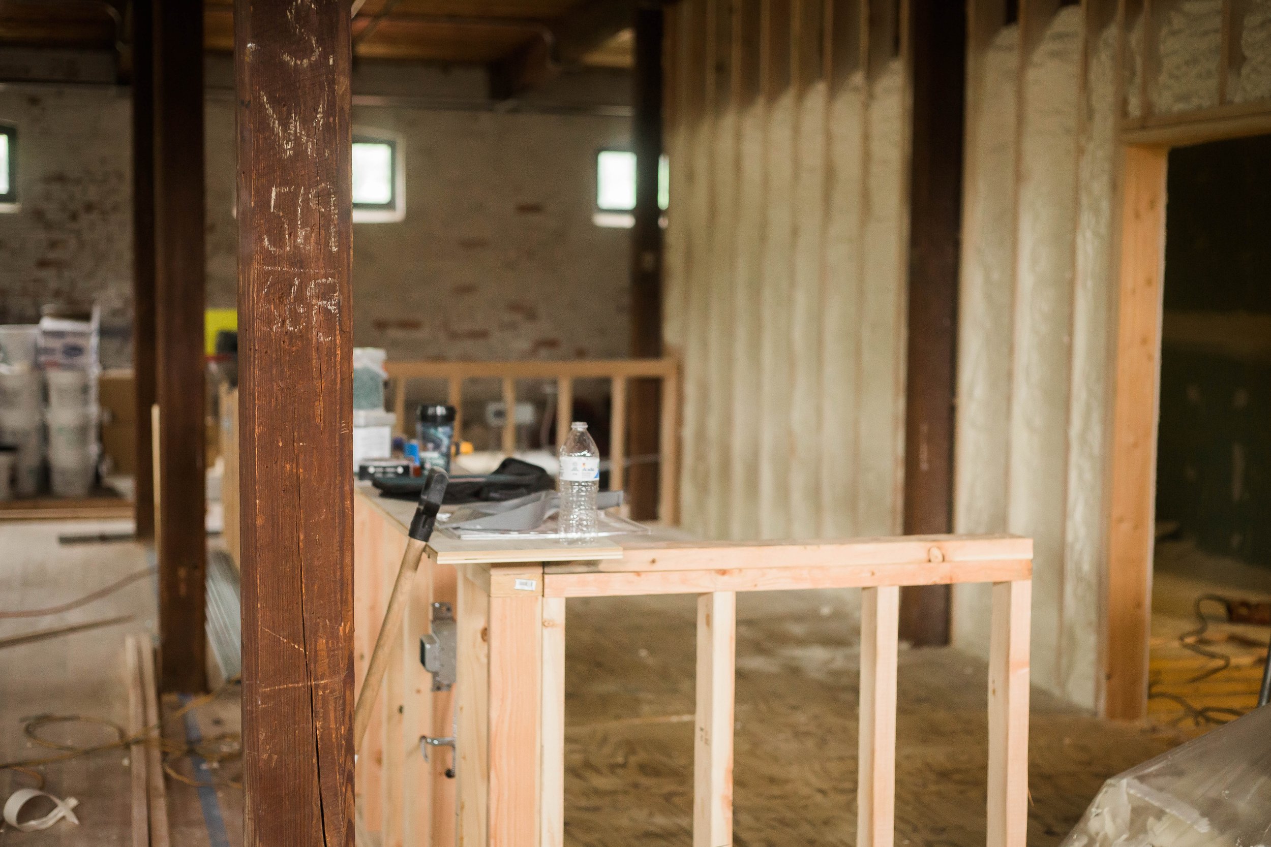 Interior view of a building under construction with wooden framing, a water bottle on a work surface, and construction tools scattered around.
