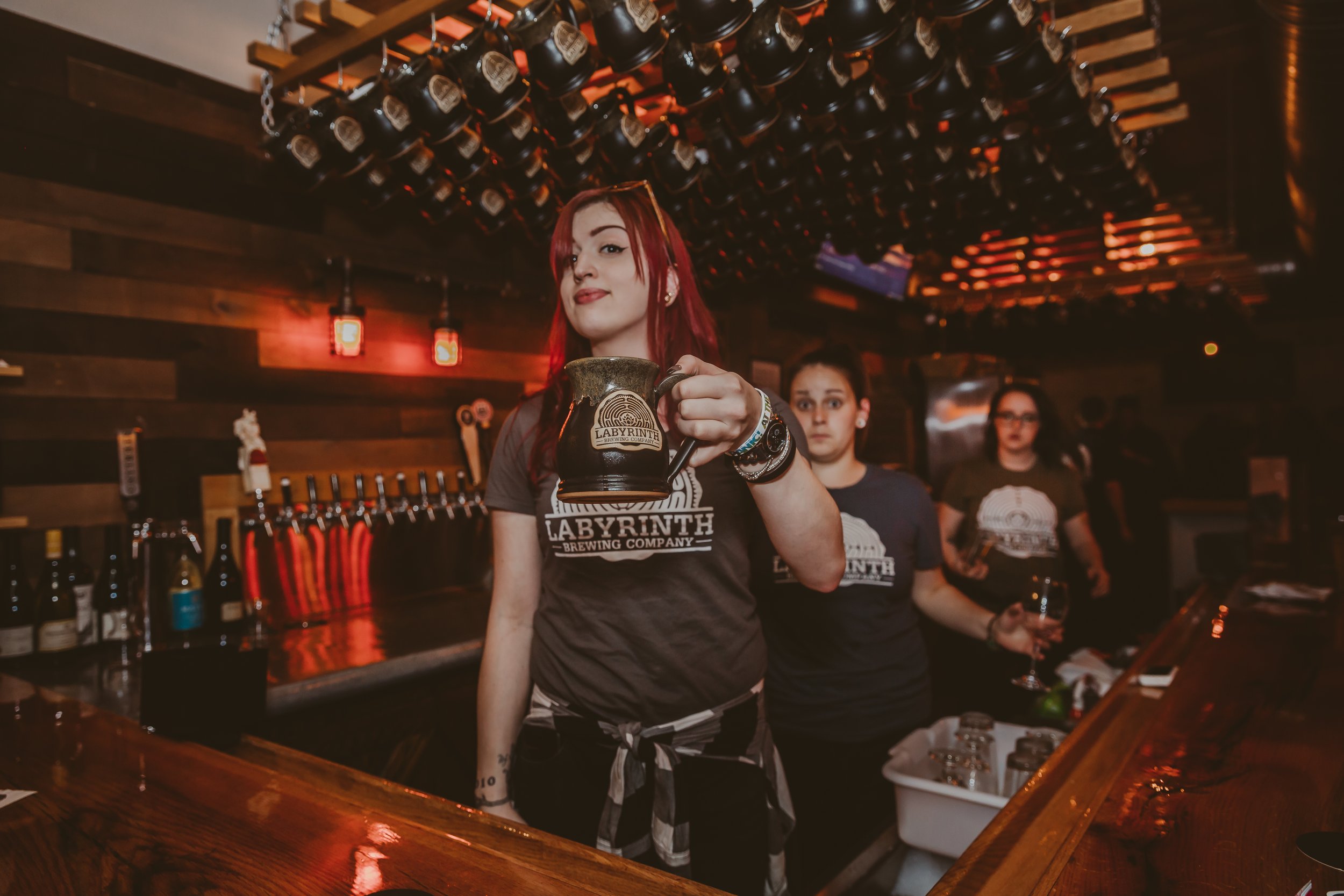A bartender with red hair holding a beer mug in a brewery bar with dark wood decor and multiple beer taps in the background.
