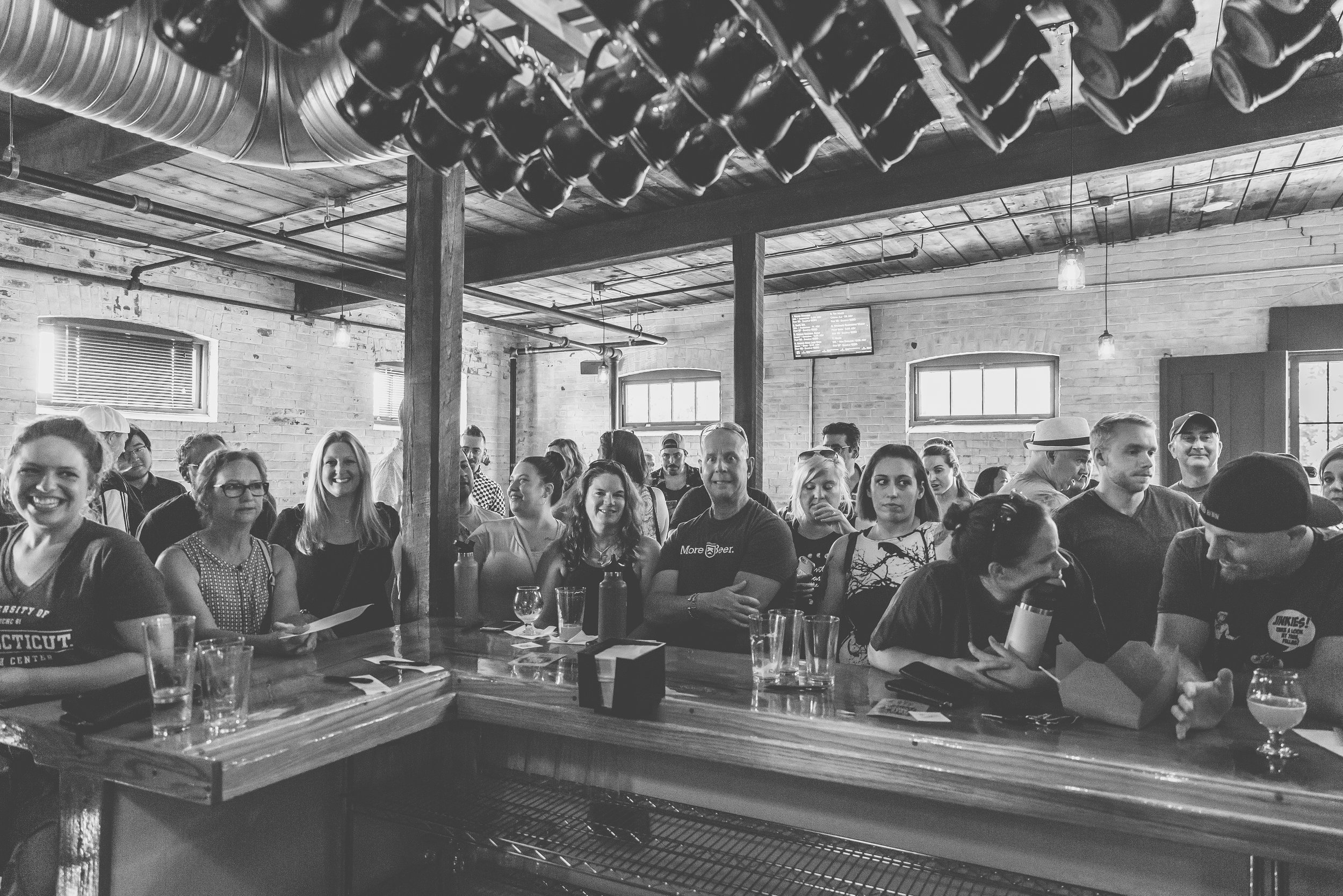 Black and white photo of a crowd of people inside a bar or pub, some smiling, some looking serious, with glasses and drink containers on the bar counter.