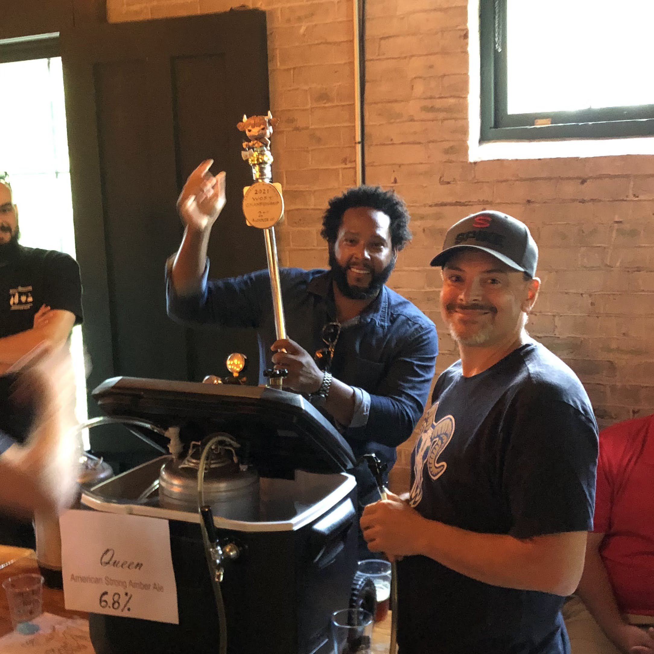 Two men at a beer tap station, one holding a tap handle with a crown and medal on top, smiling at the camera inside a brick-walled room with a window. The tap station has a sign indicating 6.8% alcohol for the American Strong Amber Ale named Queen.