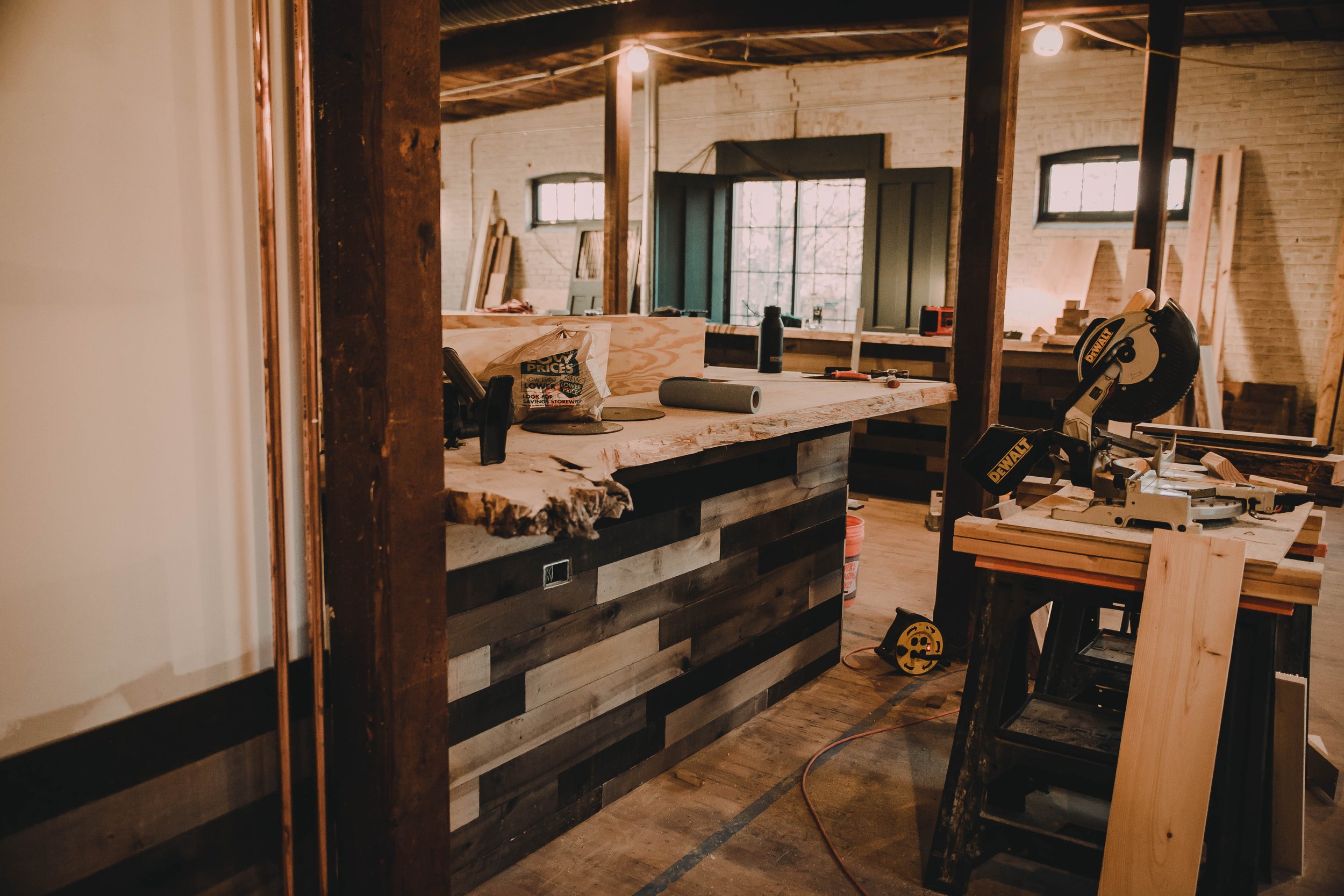 Interior of a woodworking workshop with tools, wooden planks, and workbenches in a rustic space with brick walls and large windows.