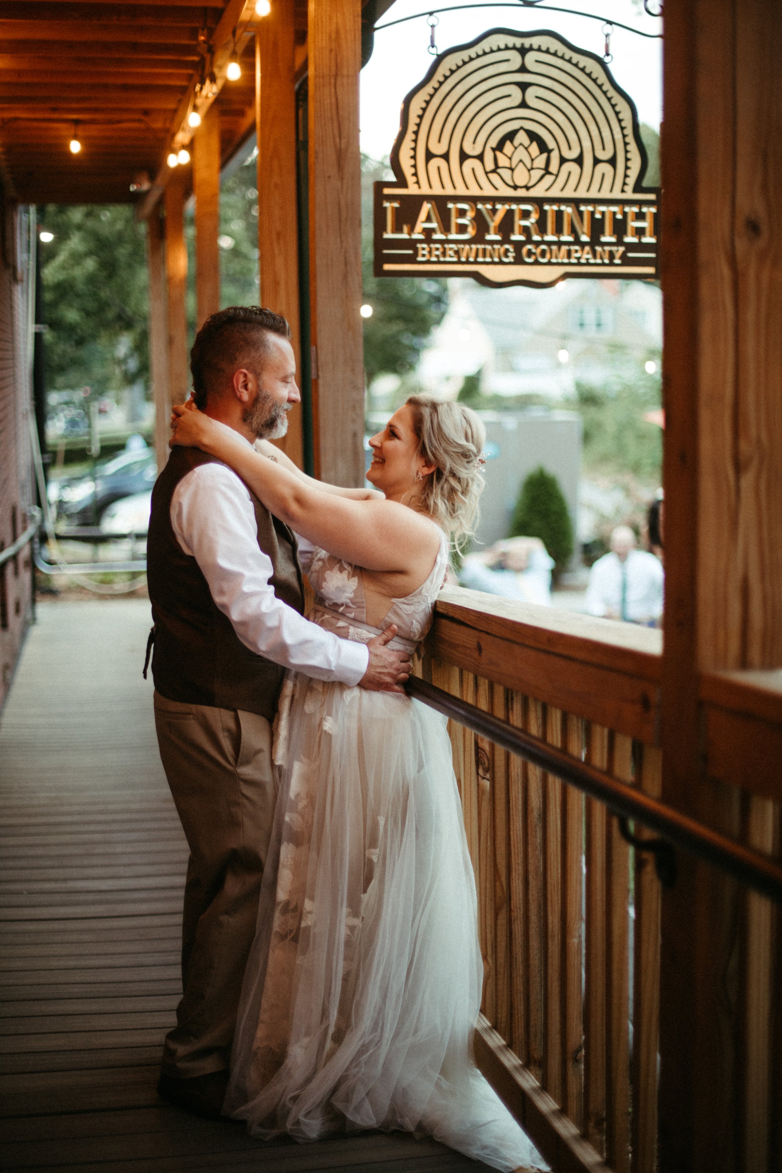 A couple, dressed in wedding attire, sharing a dance on a wooden porch outside Labyrinth Brewing Company, with a sign for the brewery overhead and trees and cars in the background.