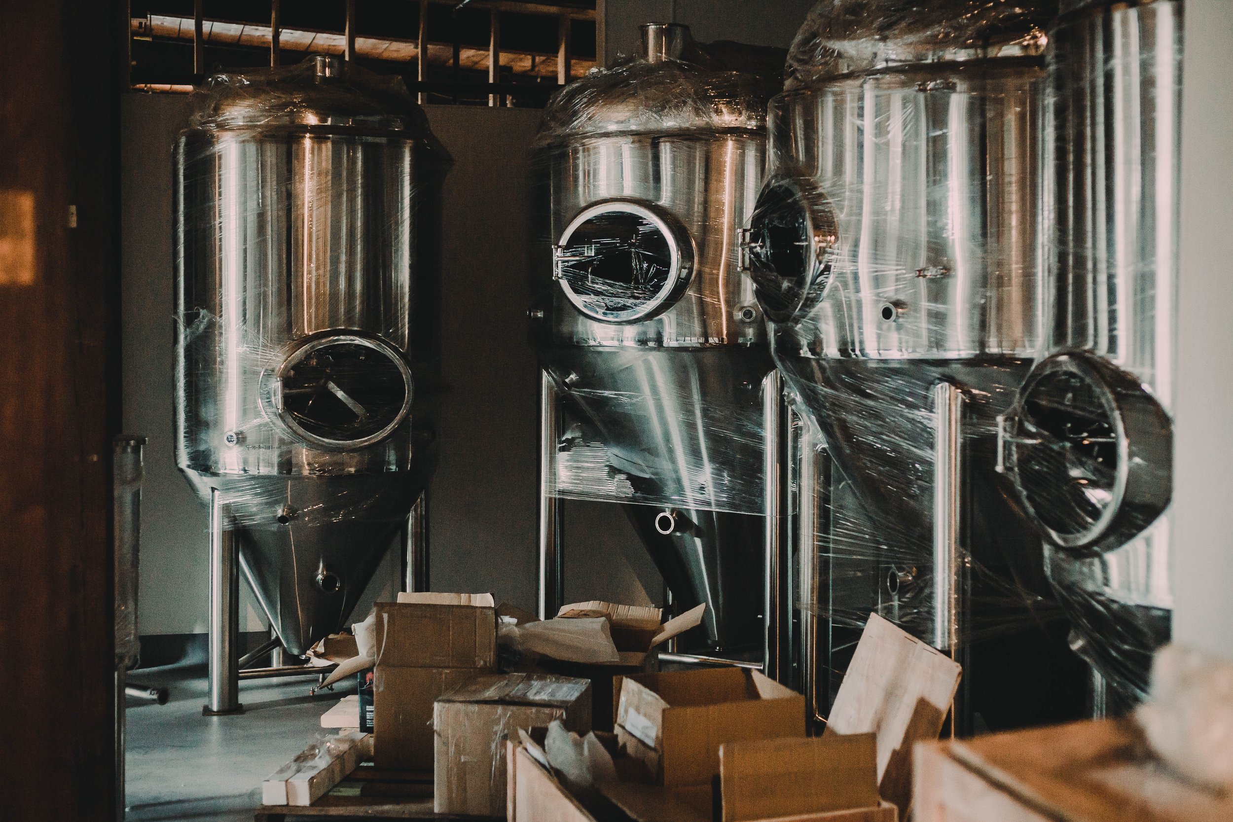 Three large stainless steel fermentation tanks in a brewery, with cardboard boxes and packing materials on the floor in front.