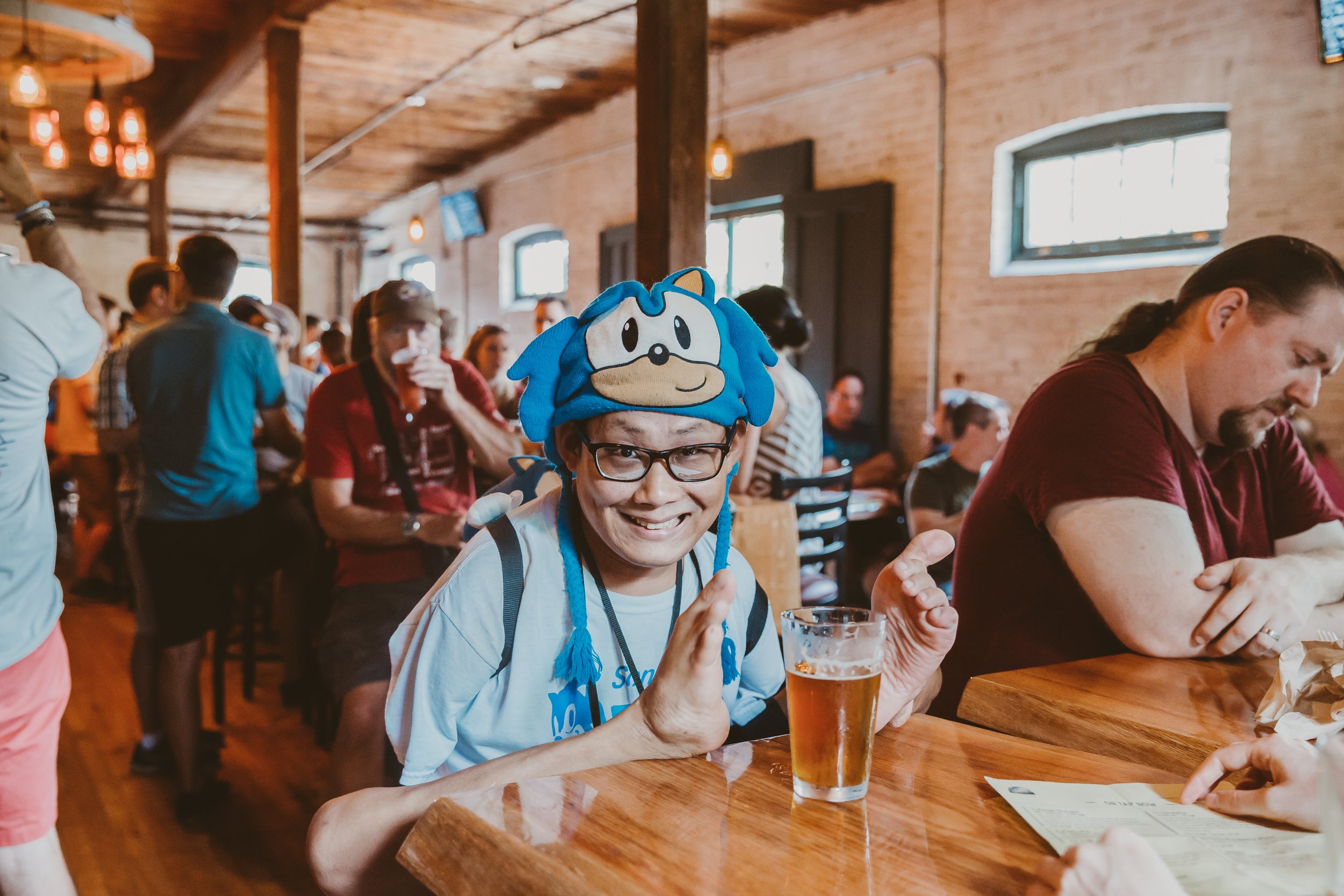 Smiling man wearing glasses and a Sonic the Hedgehog hat sitting at a bar with a drink, in a busy restaurant with brick walls and hanging lights.