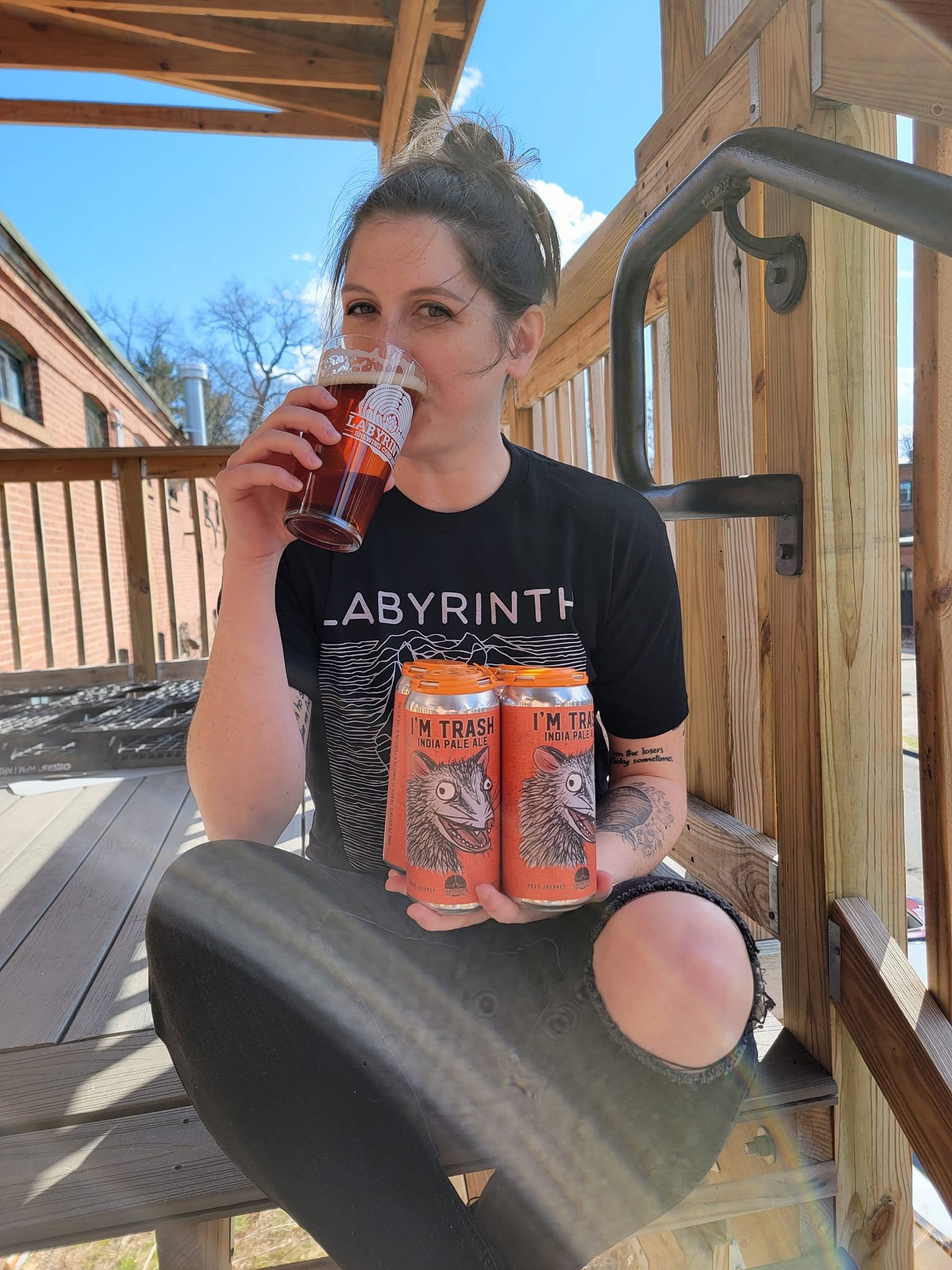 A young woman with a tattoo on her arm, sitting on a wooden outdoor staircase, drinking from a branded beer glass, holding three cans of 'I'm Trash India Pale Ale' with a cartoonish animal face on them, and smiling at the camera. She is wearing a bla