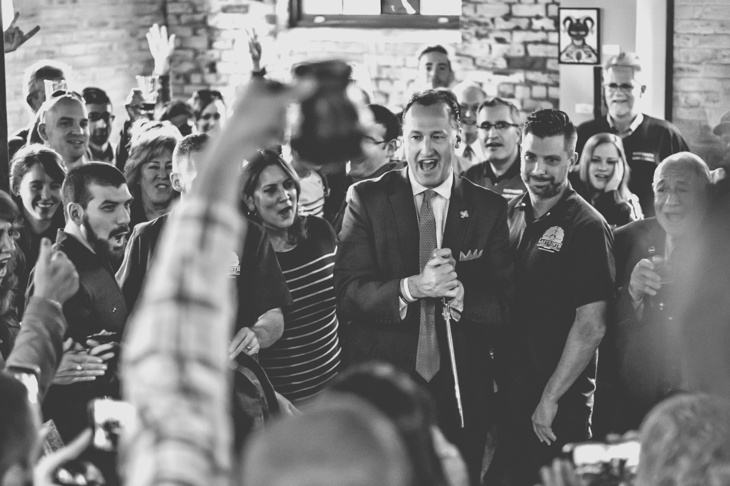 A man in a suit and tie with a crutch speaking to a crowd at an indoor event, with people smiling and taking photos.