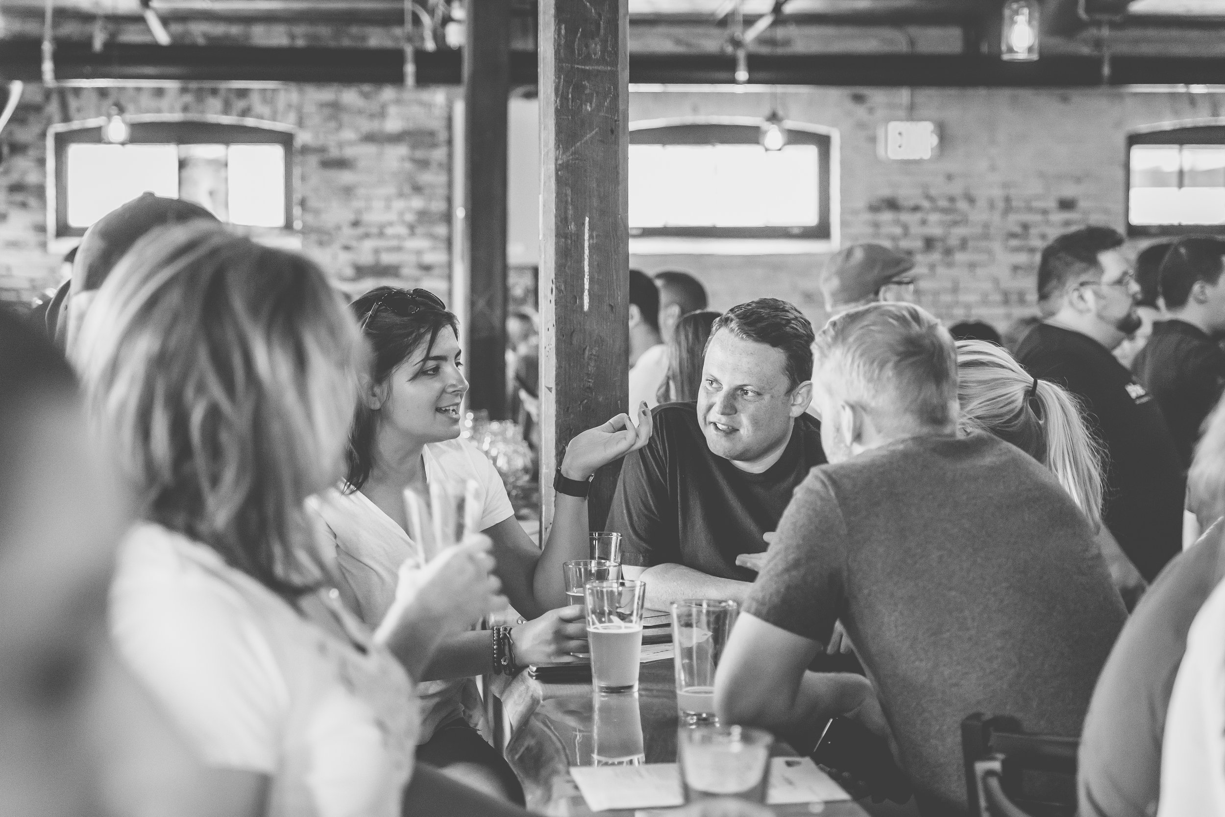 Group of people sitting at a table having conversation in a rustic bar or restaurant with exposed brick walls and large windows.