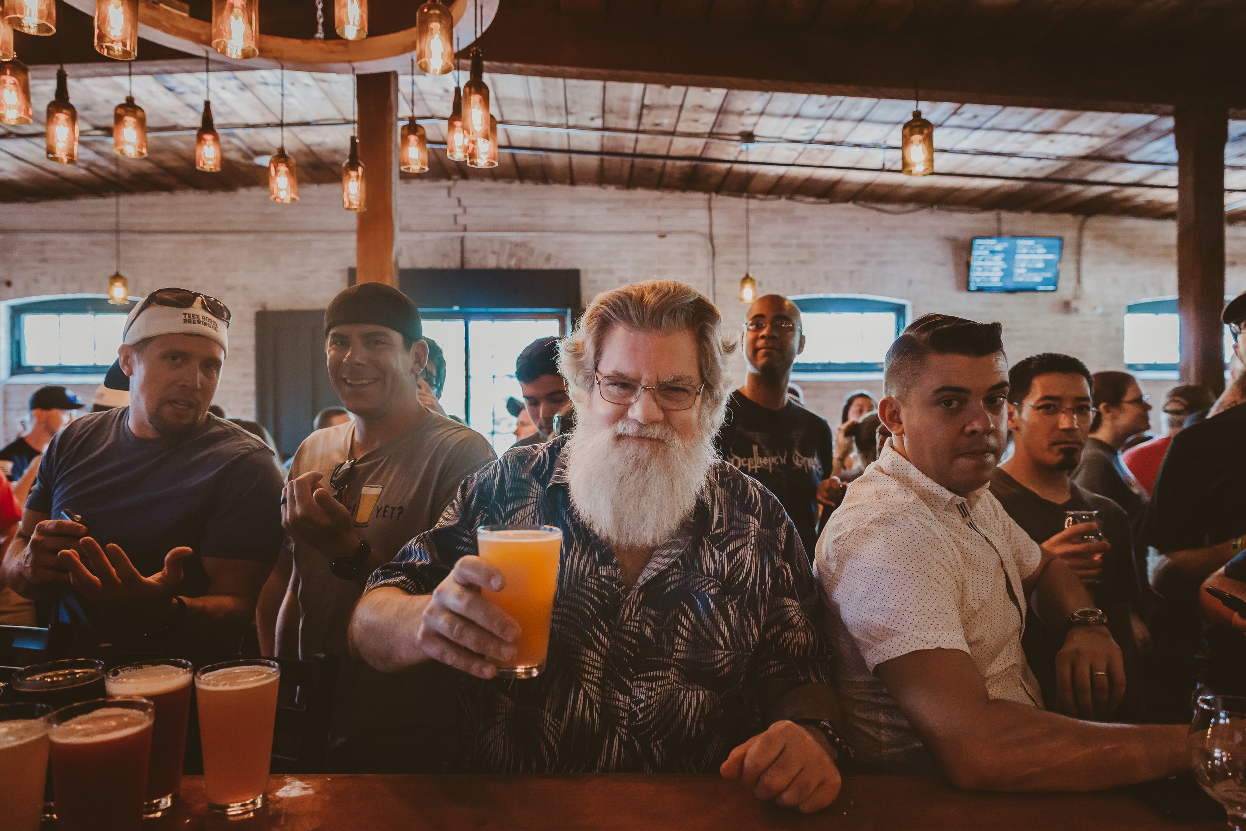 A group of people in a bar or brewery, with some holding beers, and one man with a long white beard and glasses holding a yellow drink, sitting at the bar.