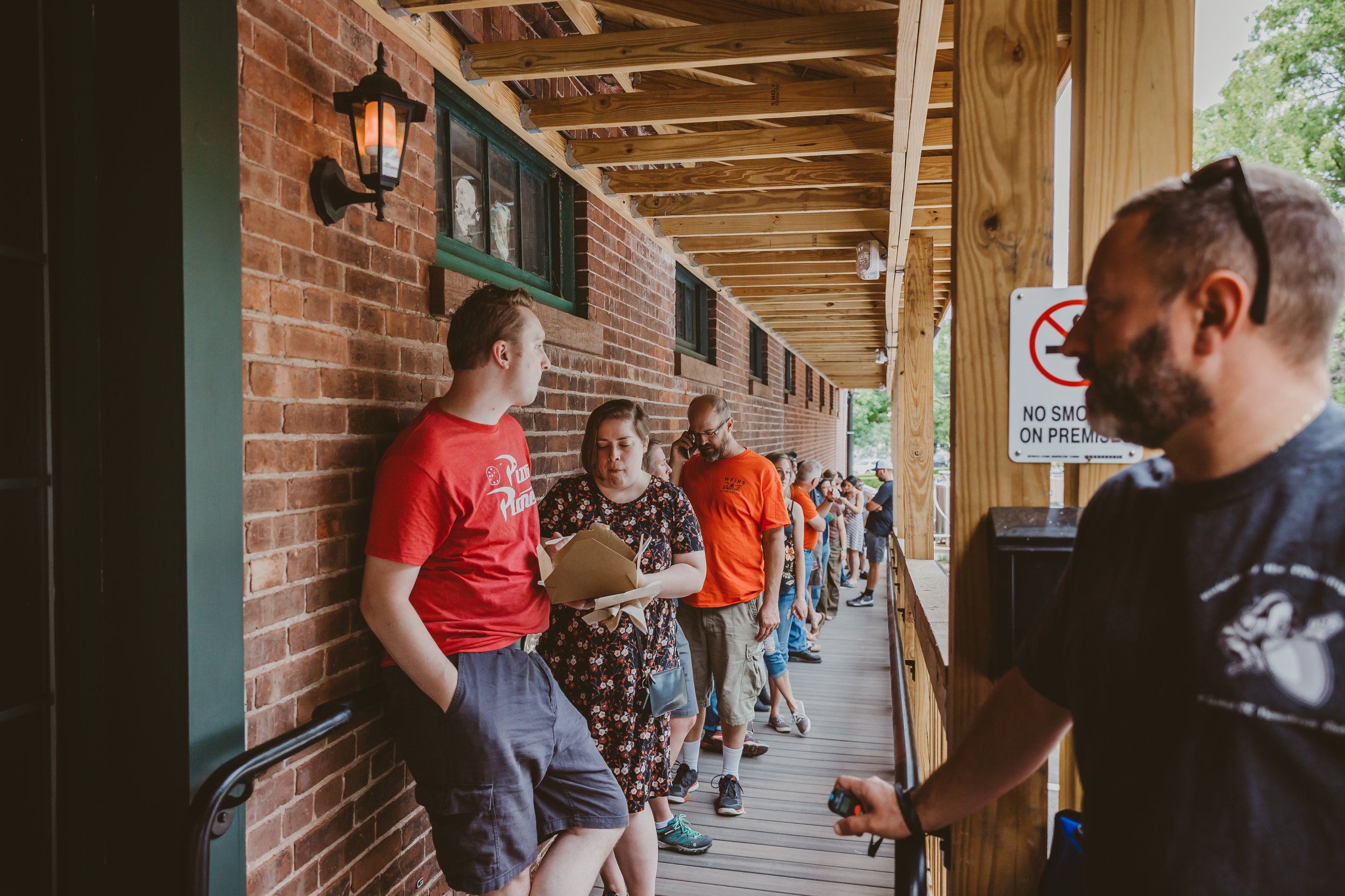 A line of people waiting outside on a wooden porch next to a brick wall, with some people looking at their phones or talking. A man in the foreground on the right is holding a device, and a woman in line is looking at her phone. There is a no smoking