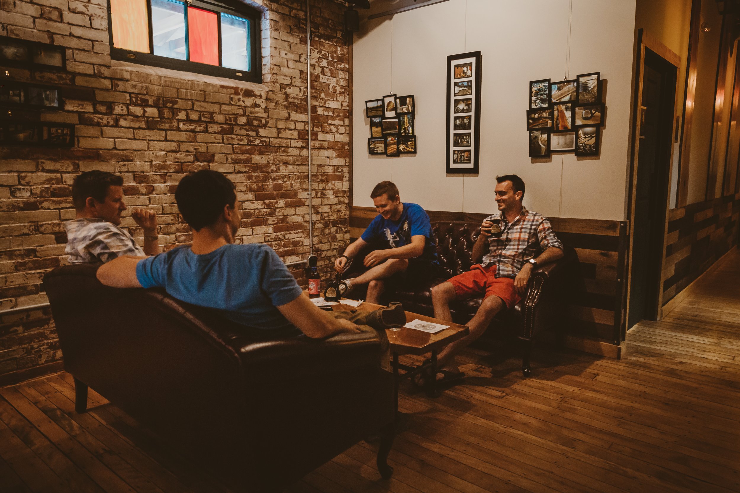 Four young men are sitting on couches in a cozy, dimly lit cafe or pub with brick and wood interior, chatting and drinking beers.