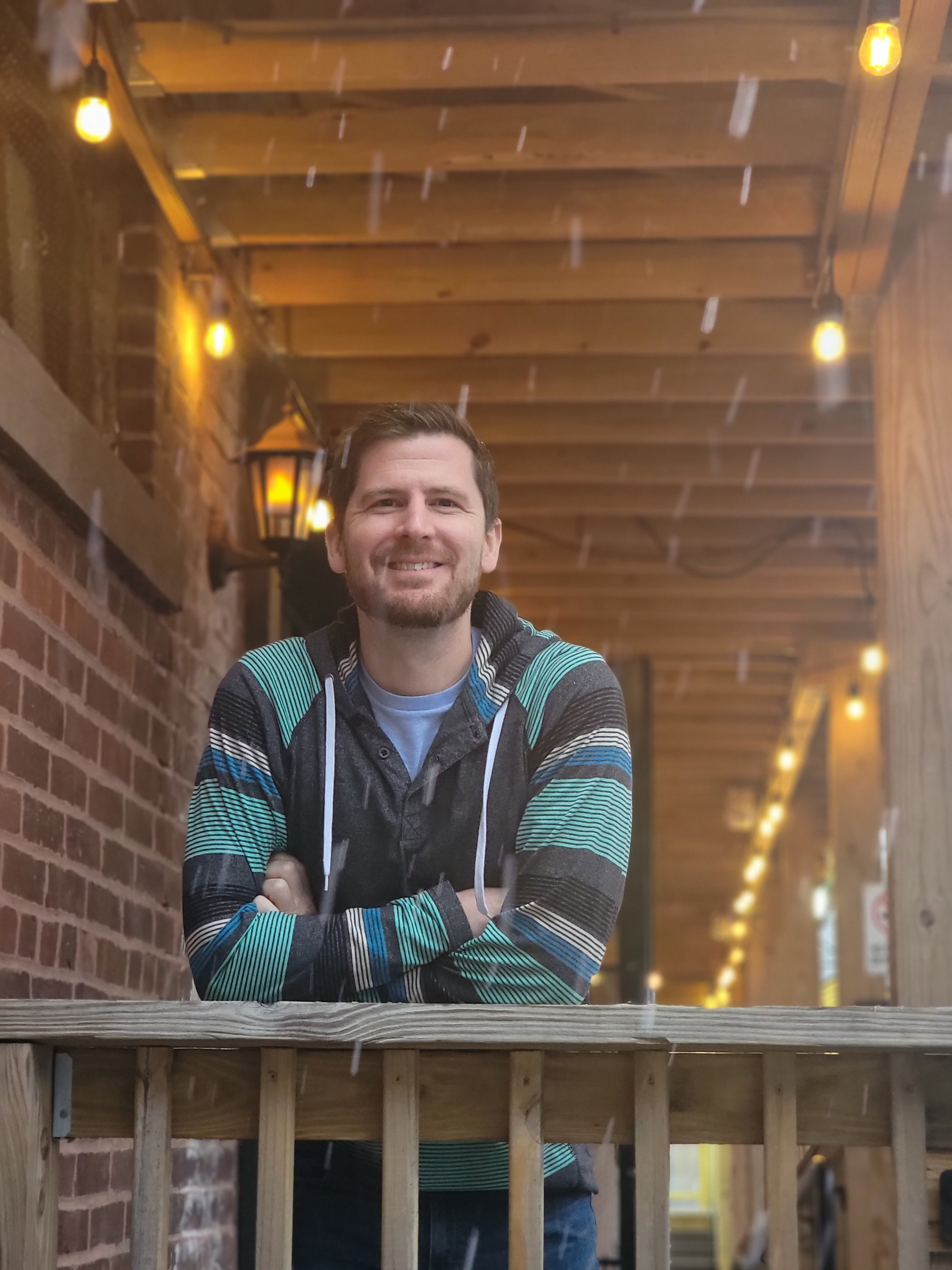 A smiling man with a beard stands outdoors under a wooden and brick shelter with string lights, wearing a striped hoodie, during rainy weather.