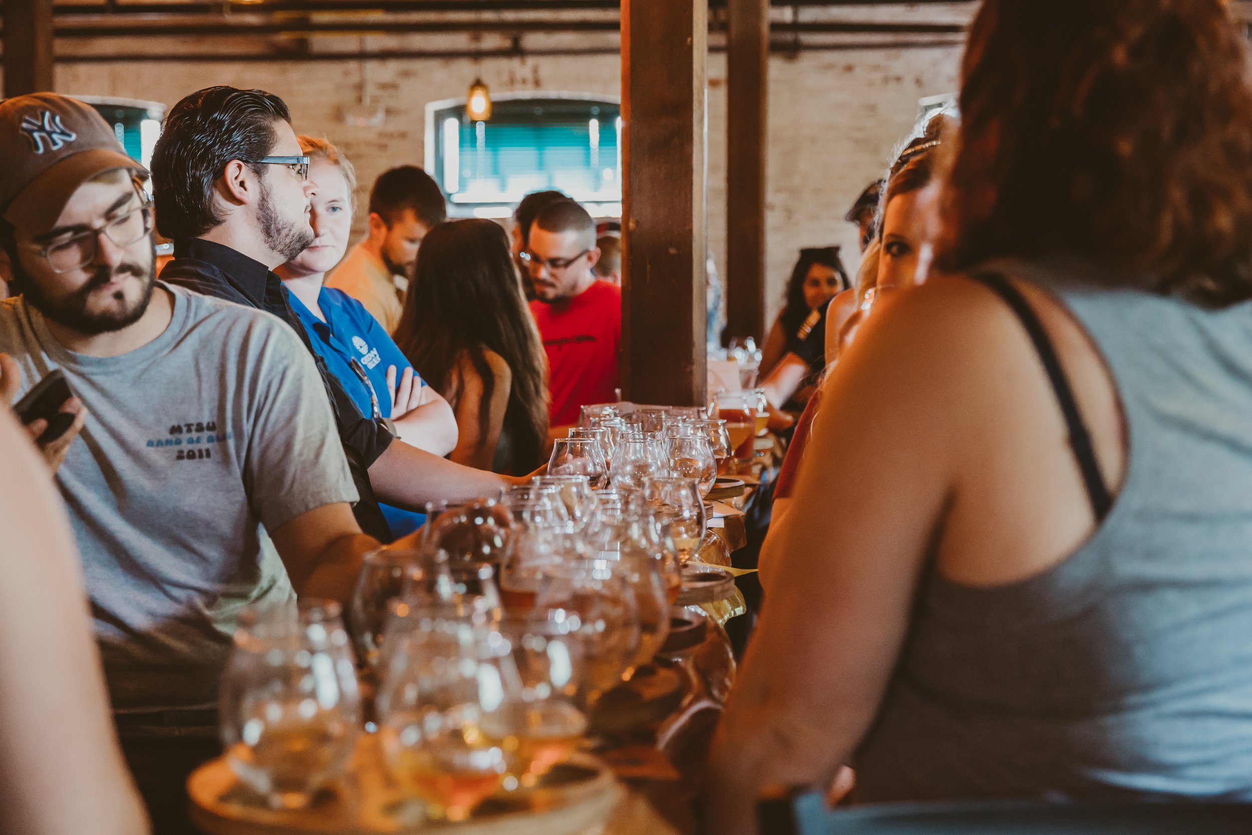 People participating in a beer tasting event, lined up at a bar with multiple glasses of beer in front of them inside a rustic venue.