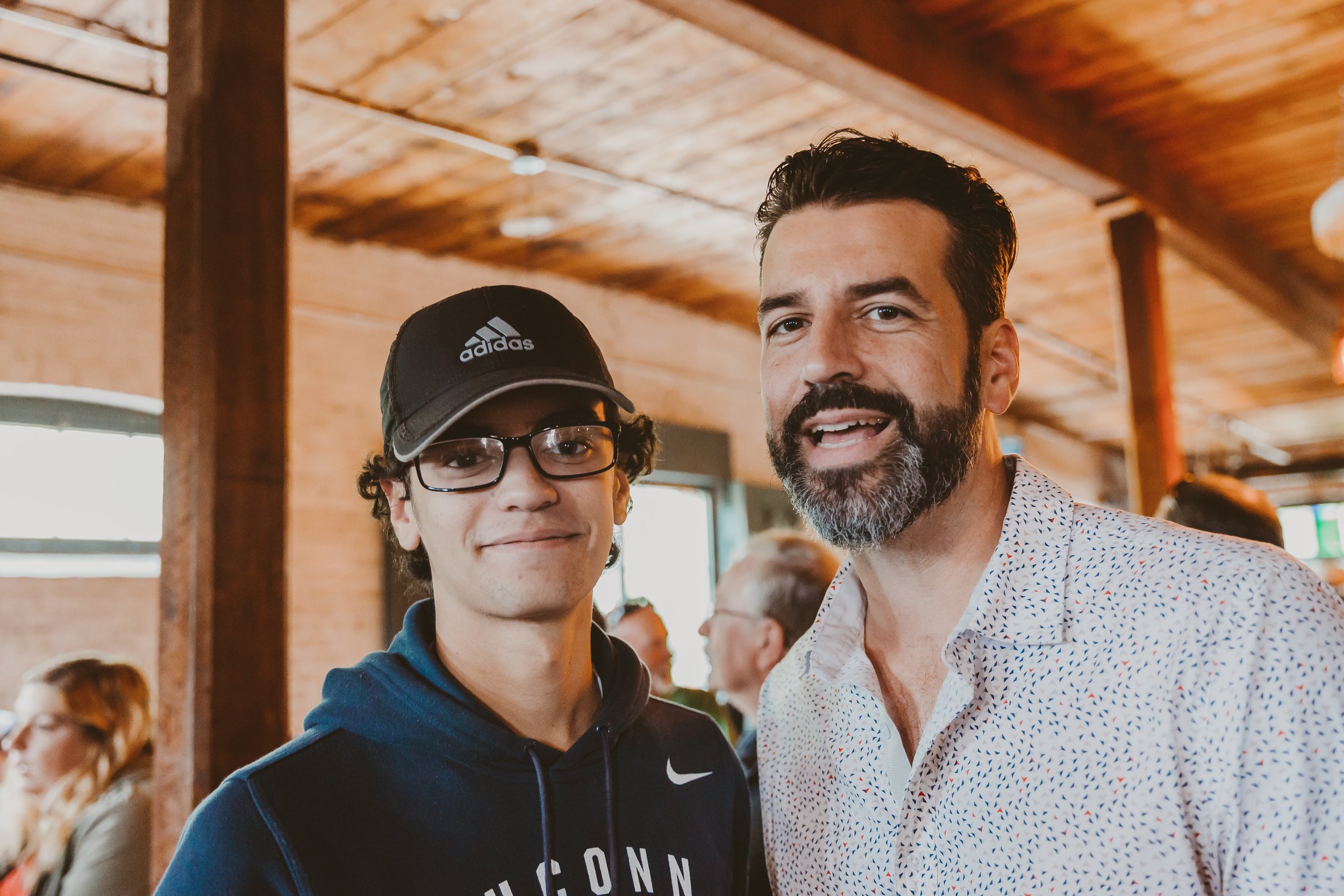 Two men, one wearing glasses and a Nike hoodie and the other with a beard and a patterned button-up shirt, smiling indoors with a wooden ceiling and other people in the background.