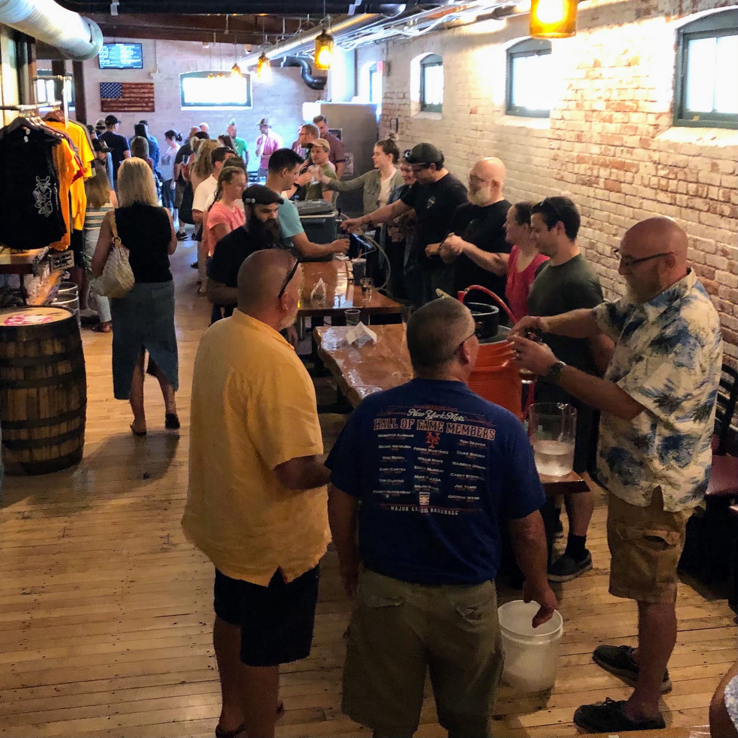 People lined up at a bar in a brewery, with some pouring drinks and others waiting. The setting has wooden floors, exposed brick walls, and hanging industrial lights, with a mix of men and women socializing.