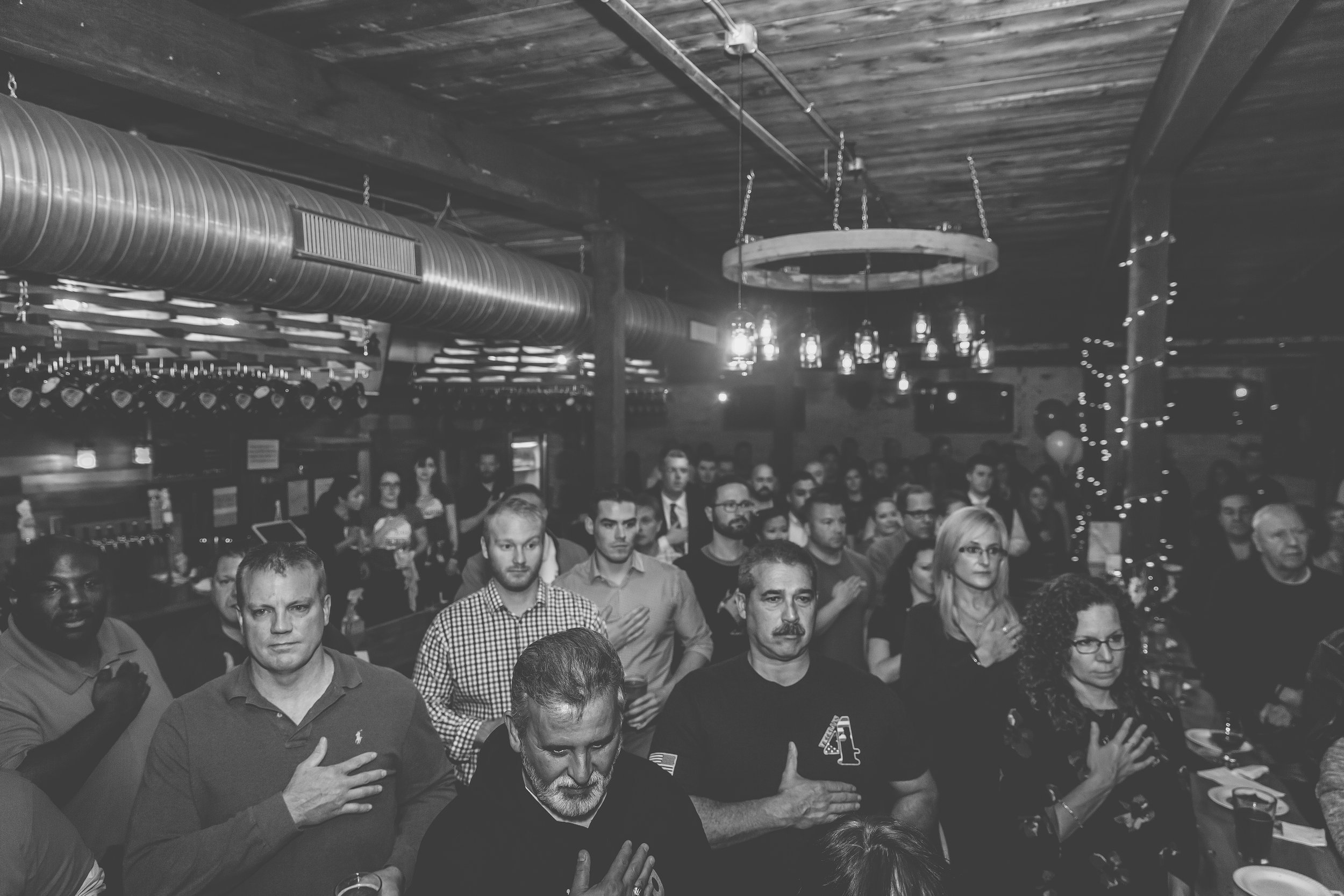 A black and white photo of a large group of diverse people standing with their right hands over their hearts in what appears to be a restaurant or bar with a wooden ceiling and hanging lights.