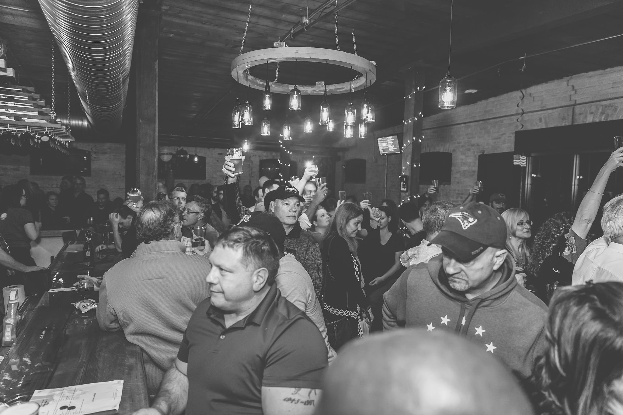Crowded bar or pub filled with people enjoying drinks and socializing. Many are raising glasses, some are talking, and others are looking around. The setting has a rustic, cozy ambiance with exposed brick walls, hanging pendant lights, and a wooden c