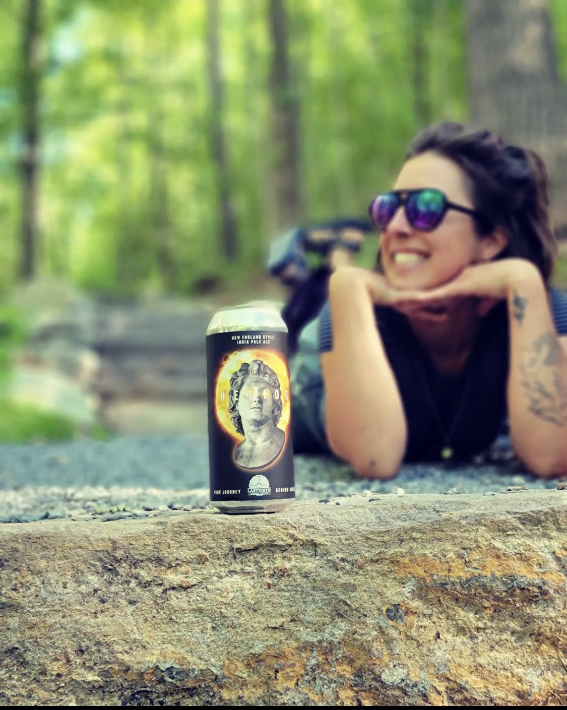 A woman lying on her stomach on a rock in a forest, smiling, wearing sunglasses, with a can of beer featuring a skull design in the foreground.