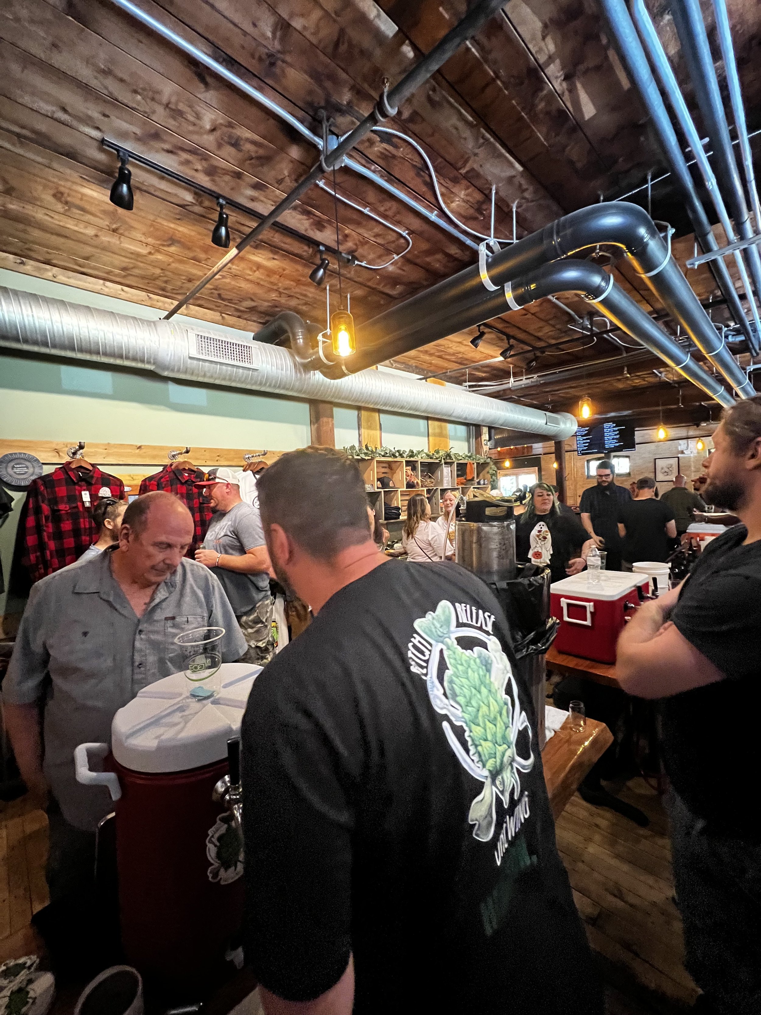 People socializing inside a rustic brewery or bar with wooden walls and ceiling, visible industrial pipes, and warm lighting.