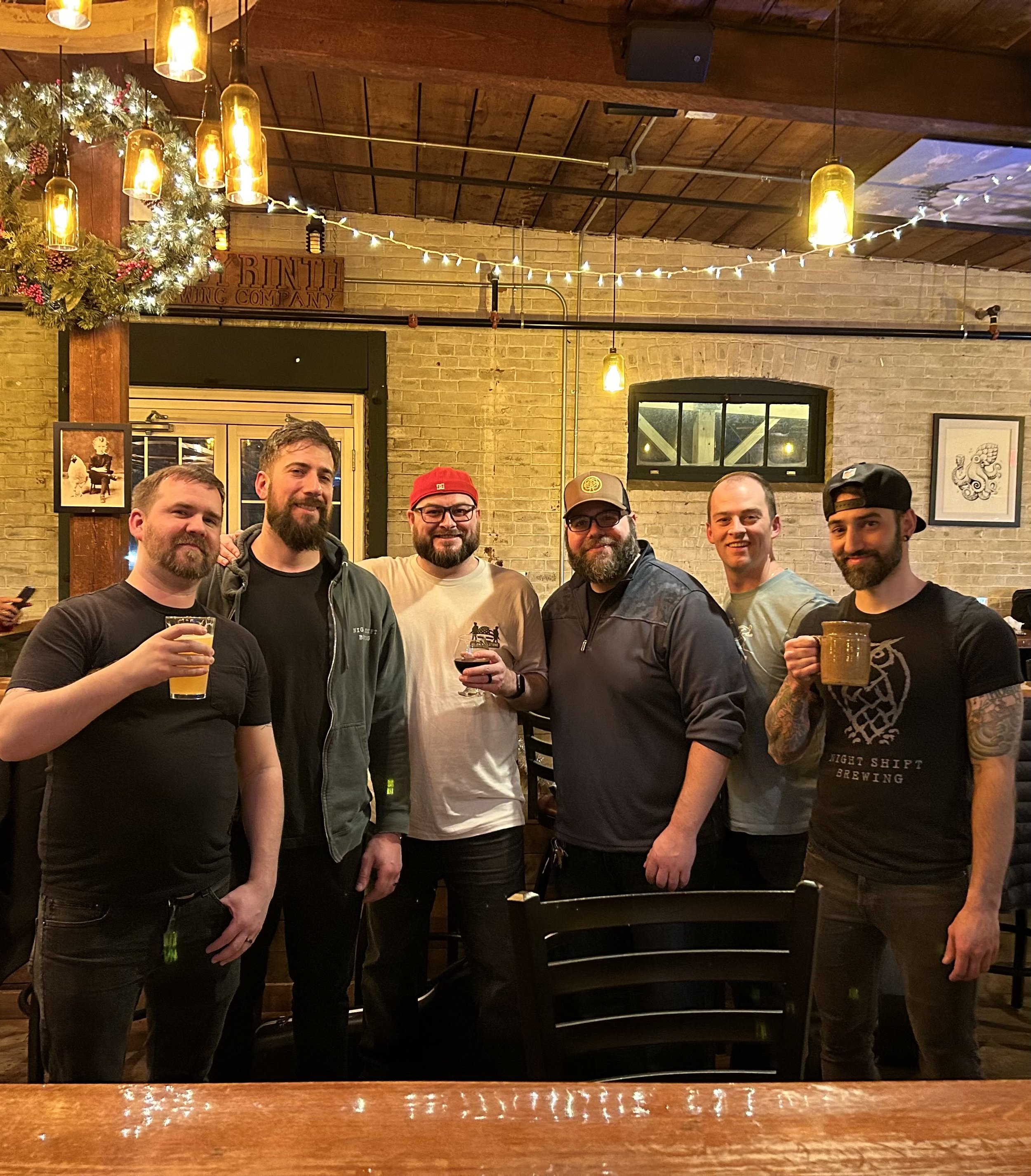 A group of six men gathered inside a cozy, warmly lit bar, holding drinks and smiling at the camera.