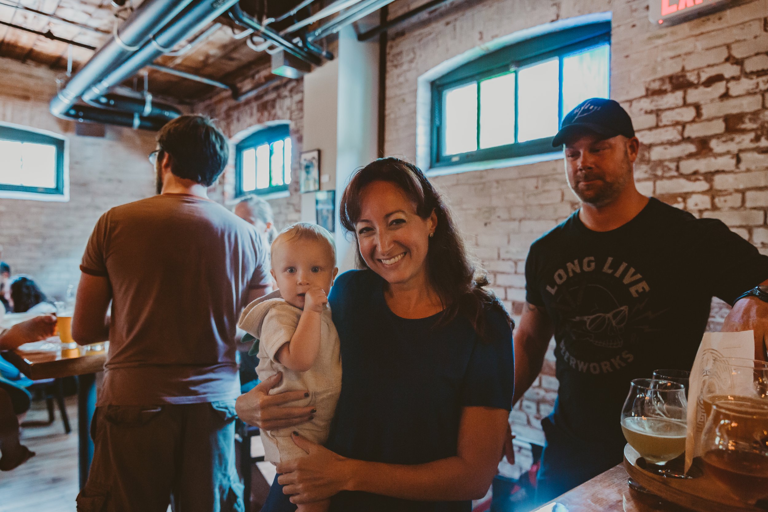 A woman holding a young child, smiling, in a bustling indoor restaurant with brick walls and large windows.