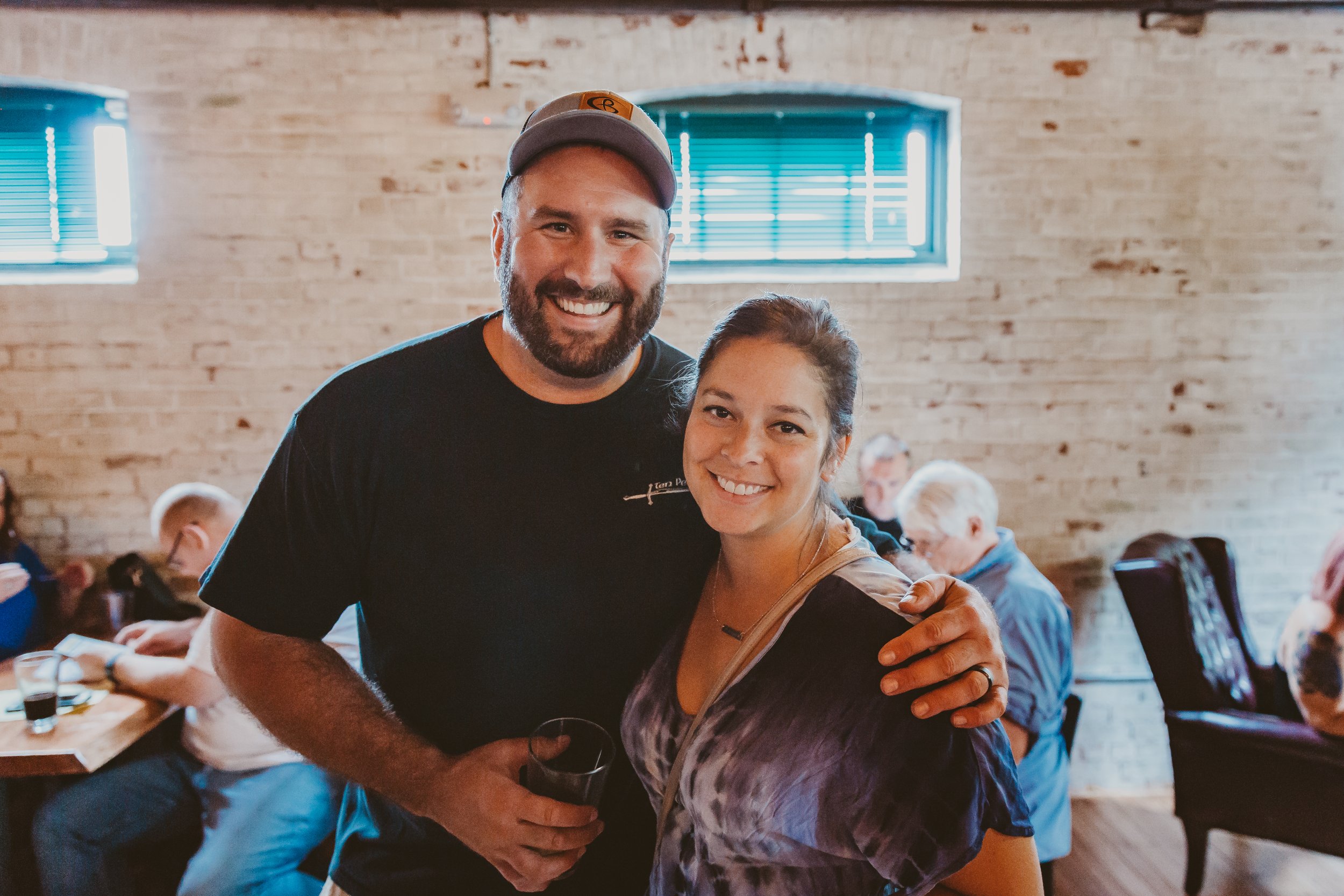 A smiling man with a beard and a woman with dark hair in a restaurant or cafe, with other people sitting at tables in the background.