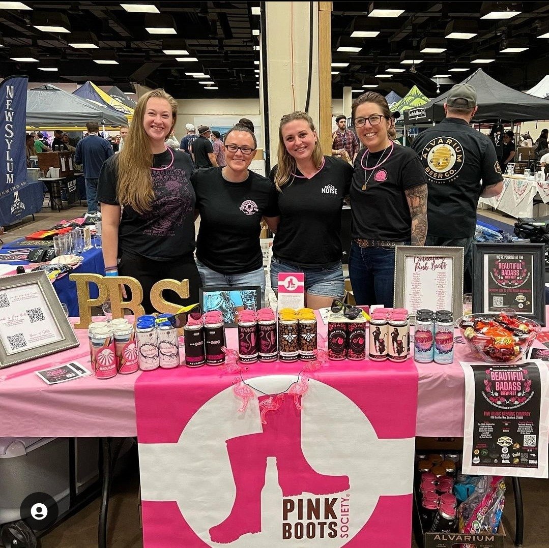 Four women standing behind a booth at a craft fair or convention, promoting Pink Boots Society. The booth is decorated with pink and black items, including cans, jars, and a large pink and white banner with a pink boot logo. The women are smiling and