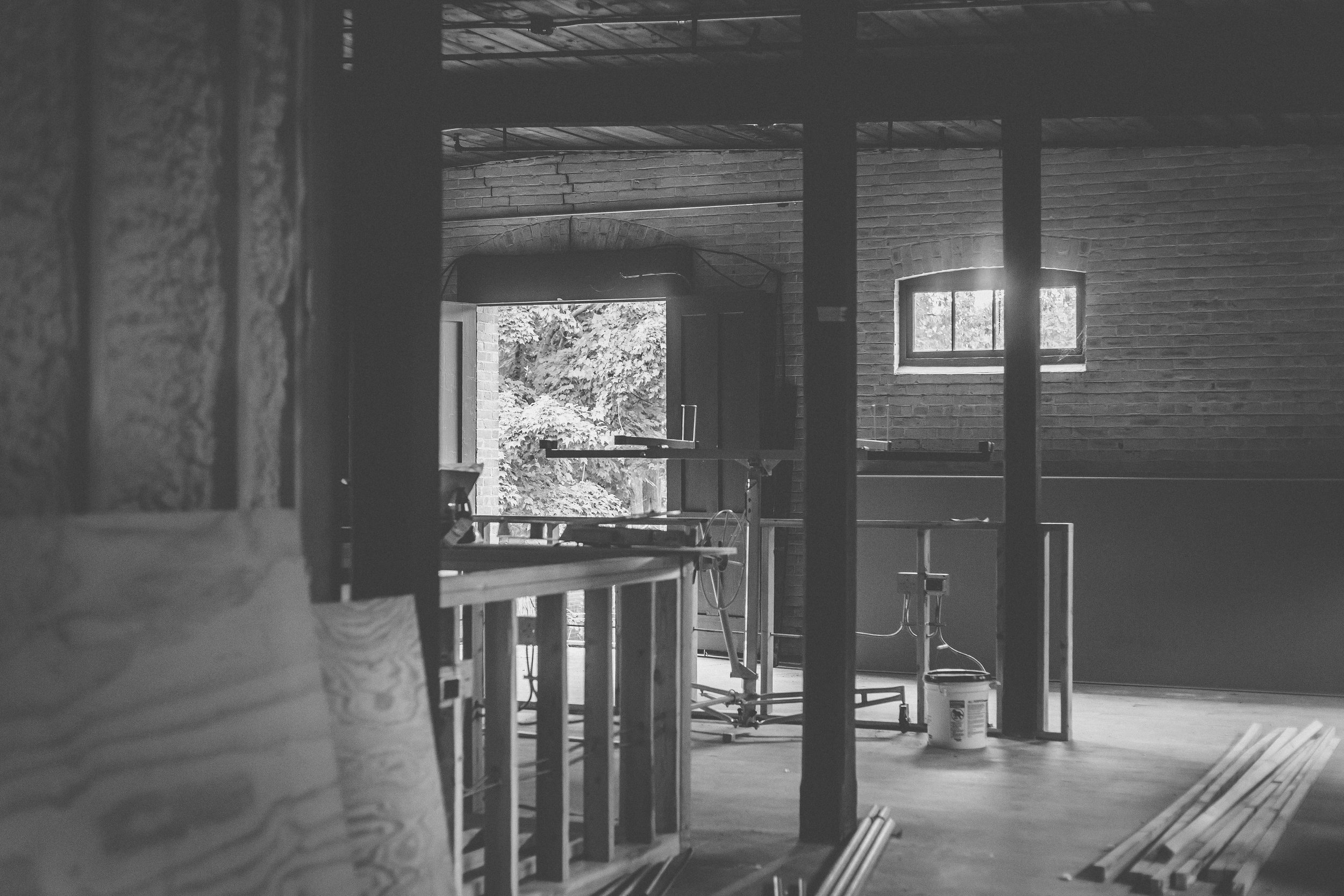 A black and white photo of an unfinished interior space with brick walls, a small window, and construction tools and materials scattered around, including a vacuum cleaner, wooden planks, and a table with equipment. Through an open door, trees and ou