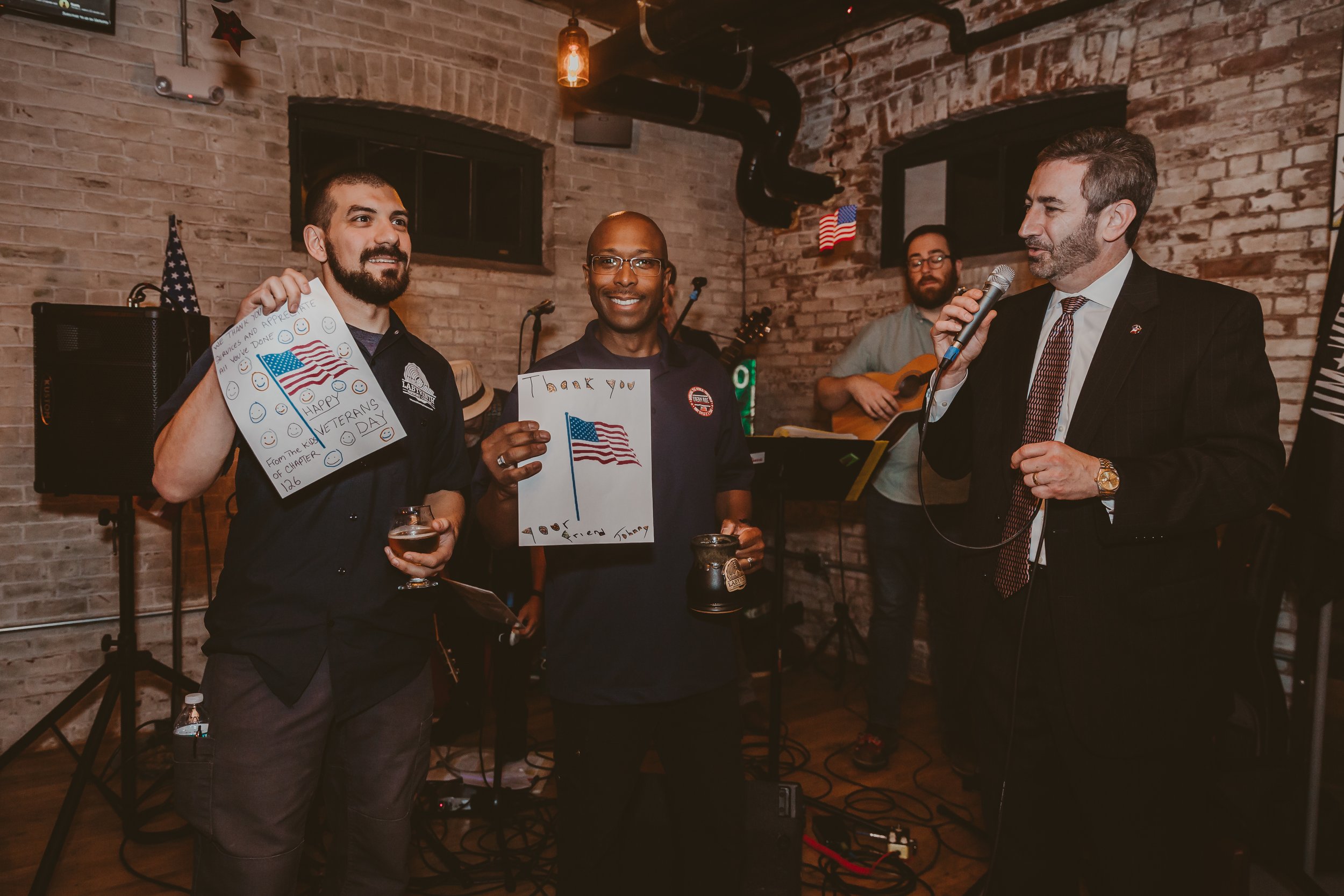 Group of men at a Veterans Day celebration, with one man singing into a microphone, two men holding handmade thank you and patriotic drawings, a musician playing guitar in the background, and American flags on the wall.