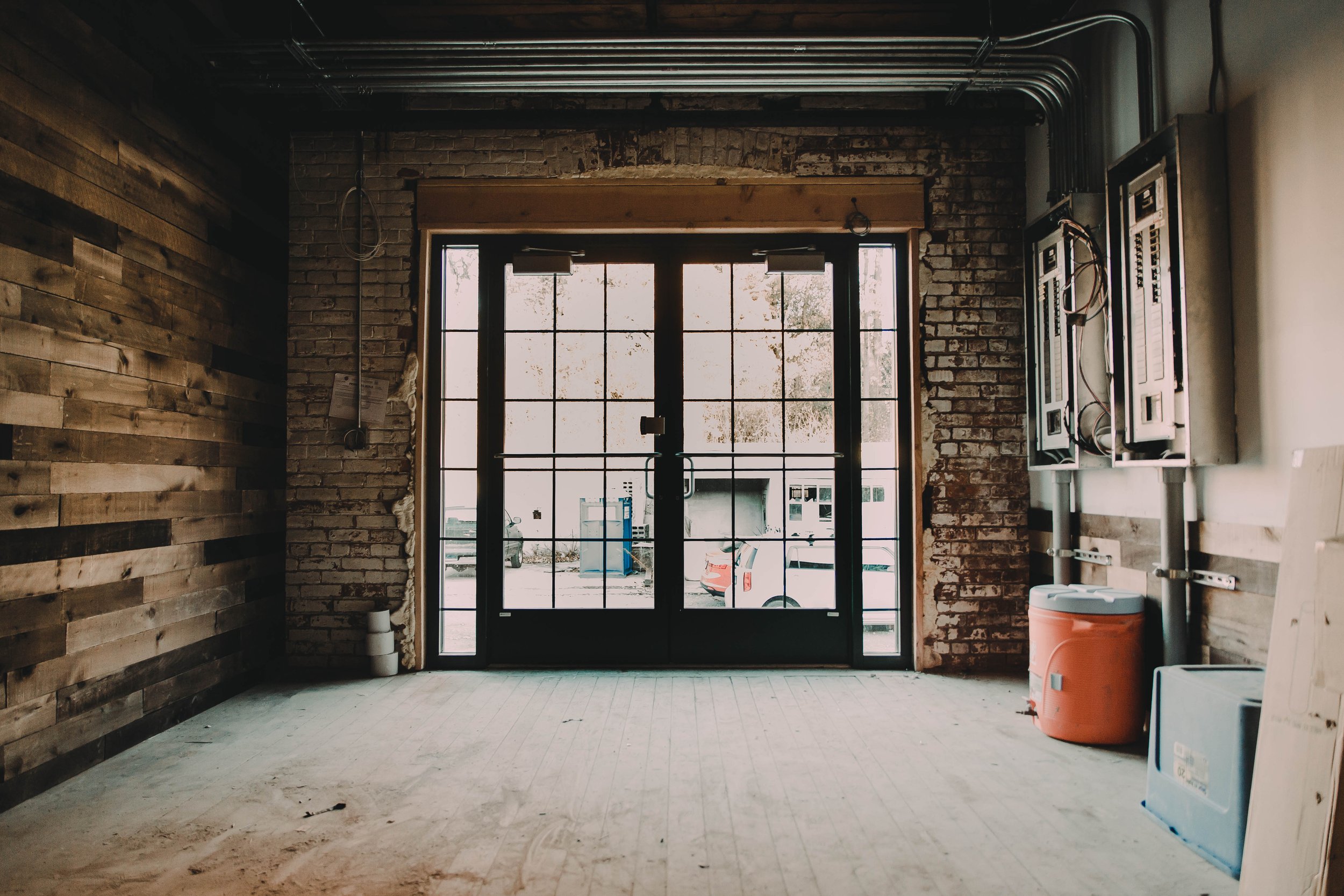 Interior of a room under construction with brick and wood walls, large glass door with grid pattern, and construction materials on the floor.