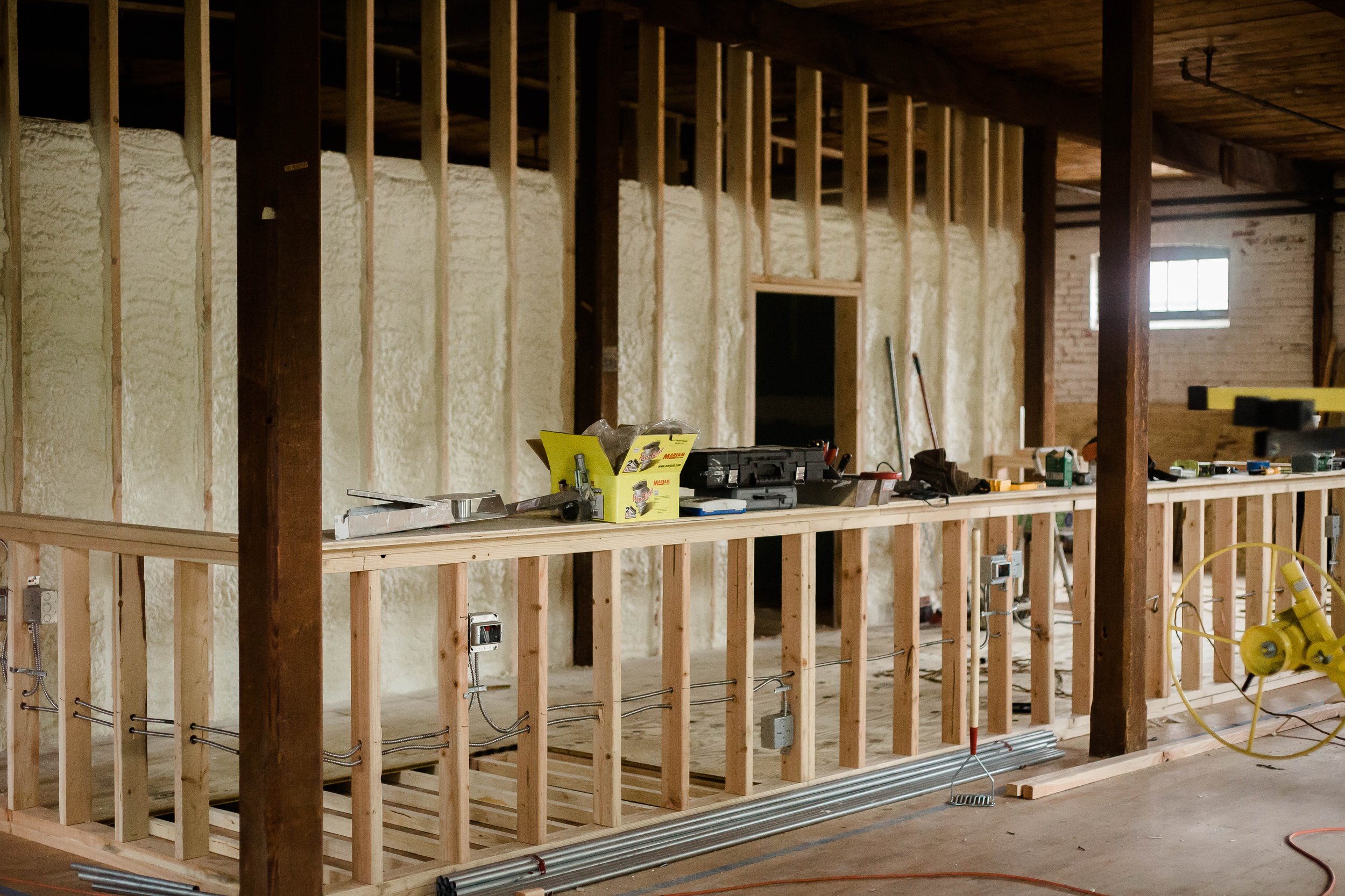 Interior of a building under construction with exposed wood framing, insulation on the walls, construction tools and materials on a work surface, and electrical wiring.
