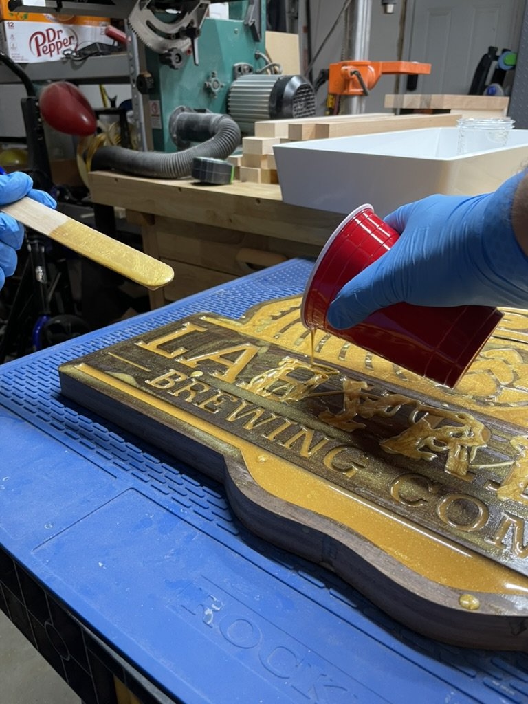 Person wearing blue gloves pouring resin onto a decorative wood sign with gold lettering that reads 'HA BREWING' in a workshop.